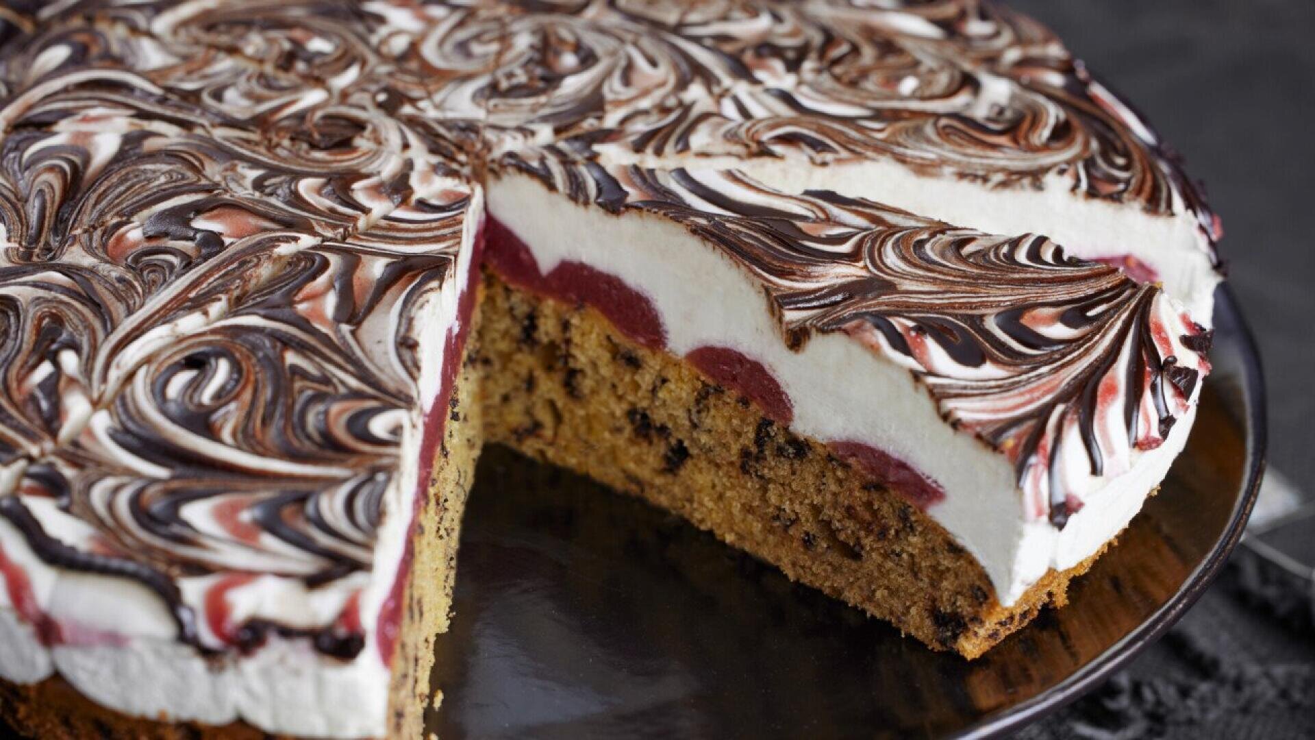 A cake with a slice removed, showing layers of chocolate chip cake, red fruit filling, and swirled white and chocolate cream on top, displayed on a dark plate.