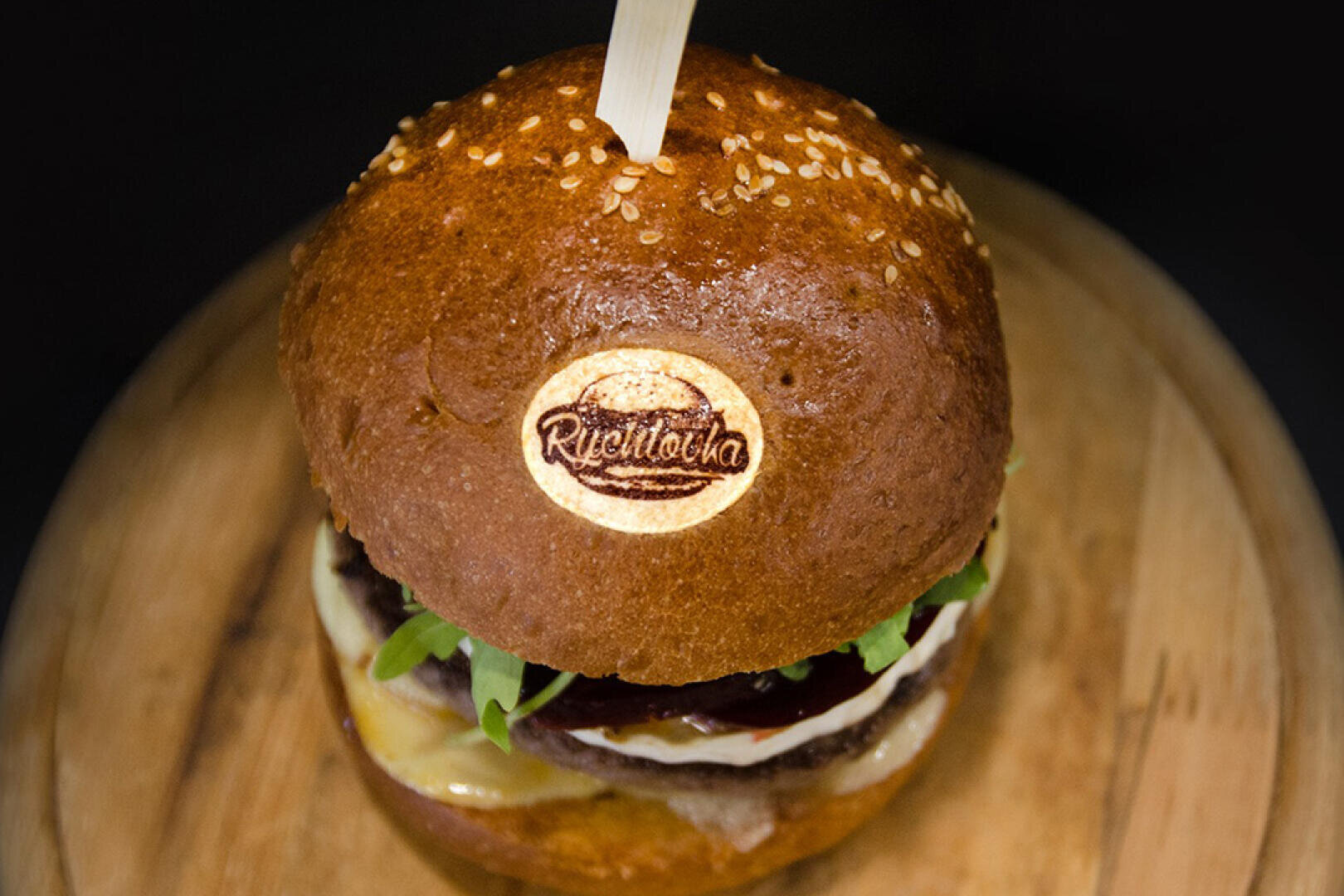 Eine Nahaufnahme eines Gourmet-Hamburger mit Sesambrötchen, Blattgemüse, geschmolzenem Käse und einem Markenlogo mit der Aufschrift Riettocha auf dem oberen Brötchen, serviert auf einem Holzbrett mit einem Spieß in der Mitte.