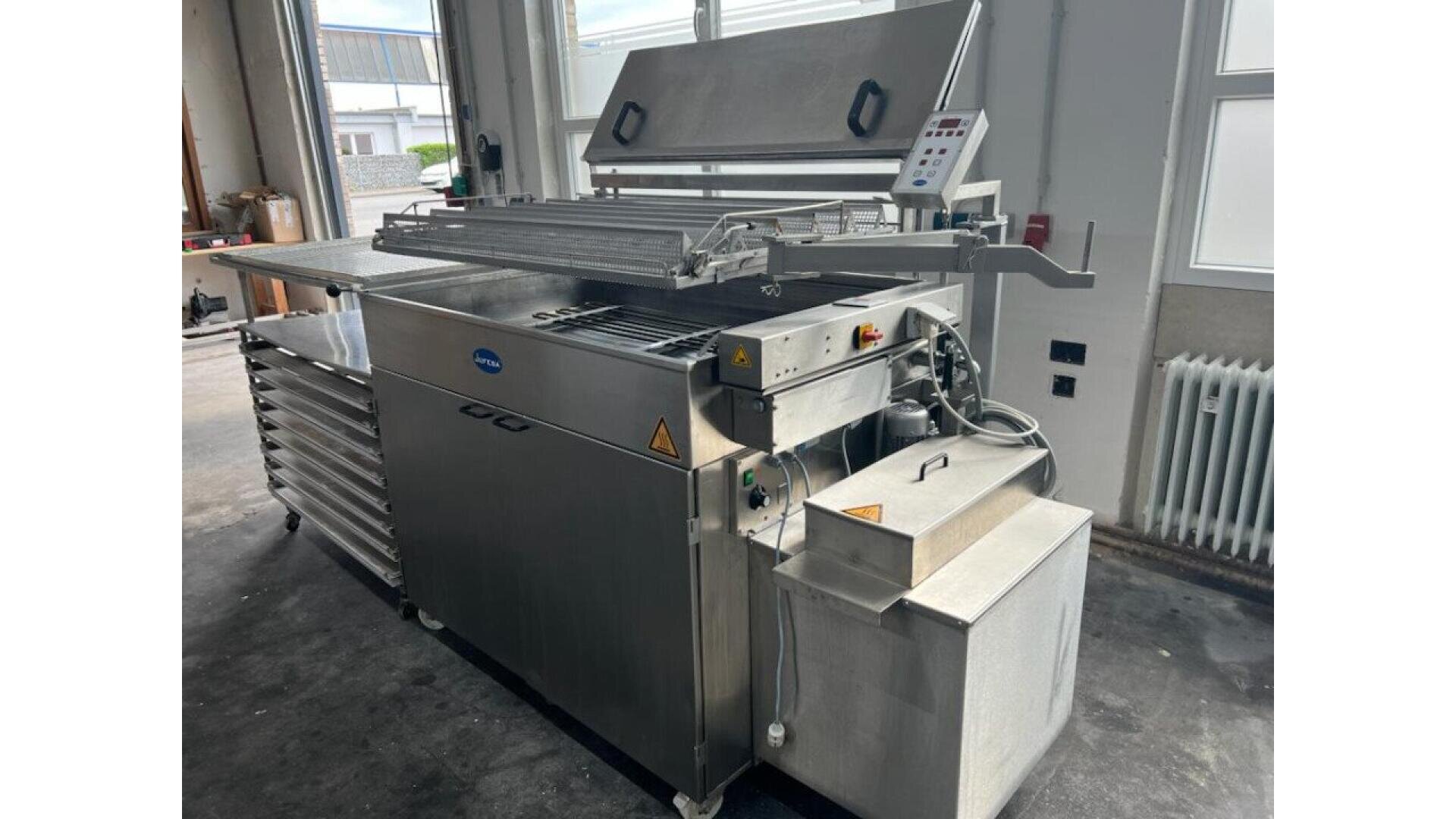 A large industrial stainless steel machine with an open lid, control panel, and various compartments stands in a workshop with concrete floors, windows, and a radiator in the background.