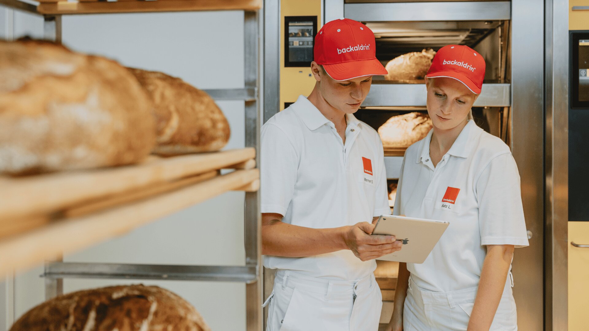 Zwei Bäcker in weißen Uniformen und roten Mützen schauen gemeinsam auf eine Tafel in einer Bäckerei, um die herum frische Brote in Regalen und Öfen liegen.
