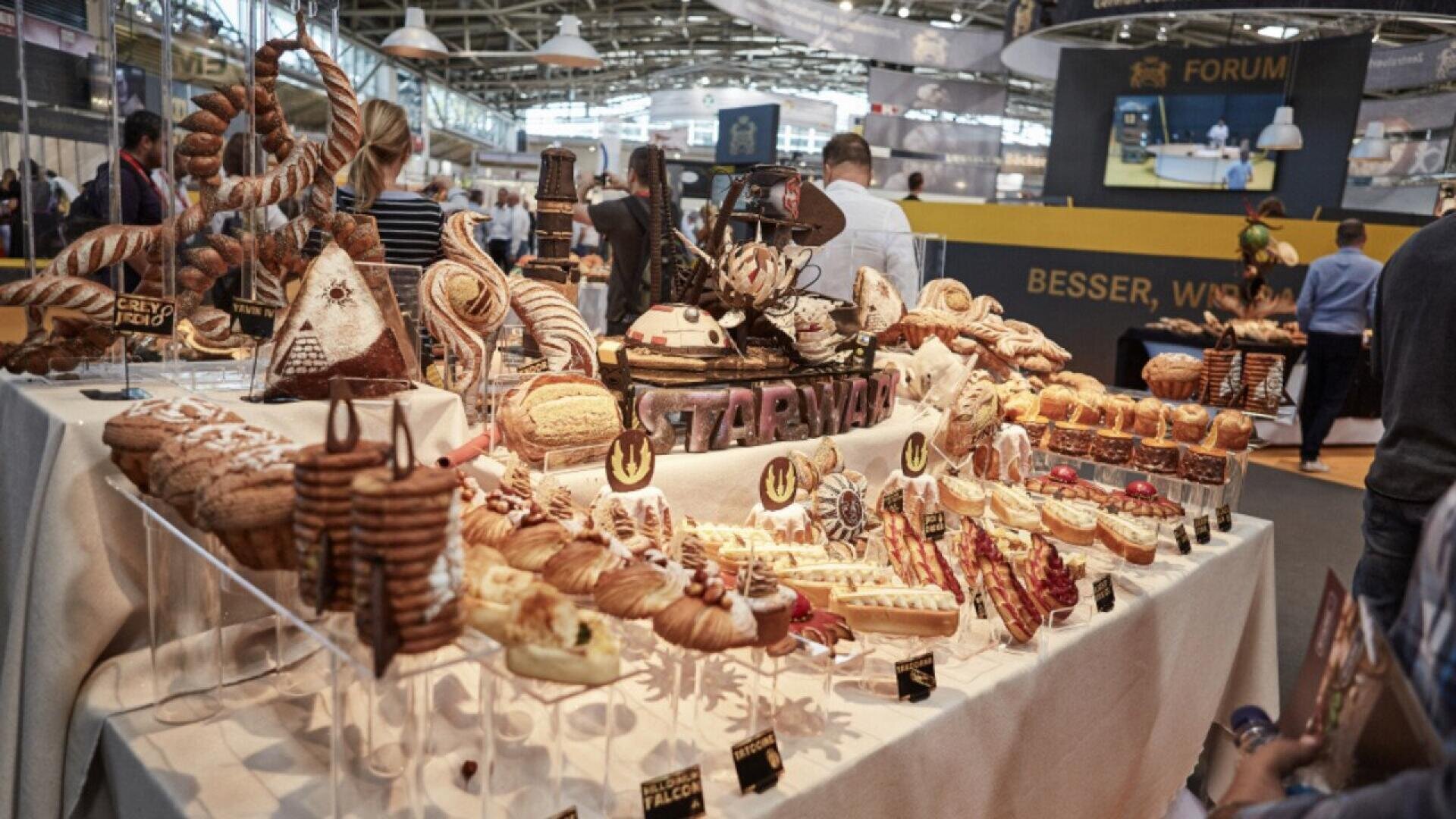A table covered with a variety of intricately arranged breads and pastries is displayed at a busy indoor food exhibition, with people browsing and tasting in the background.
