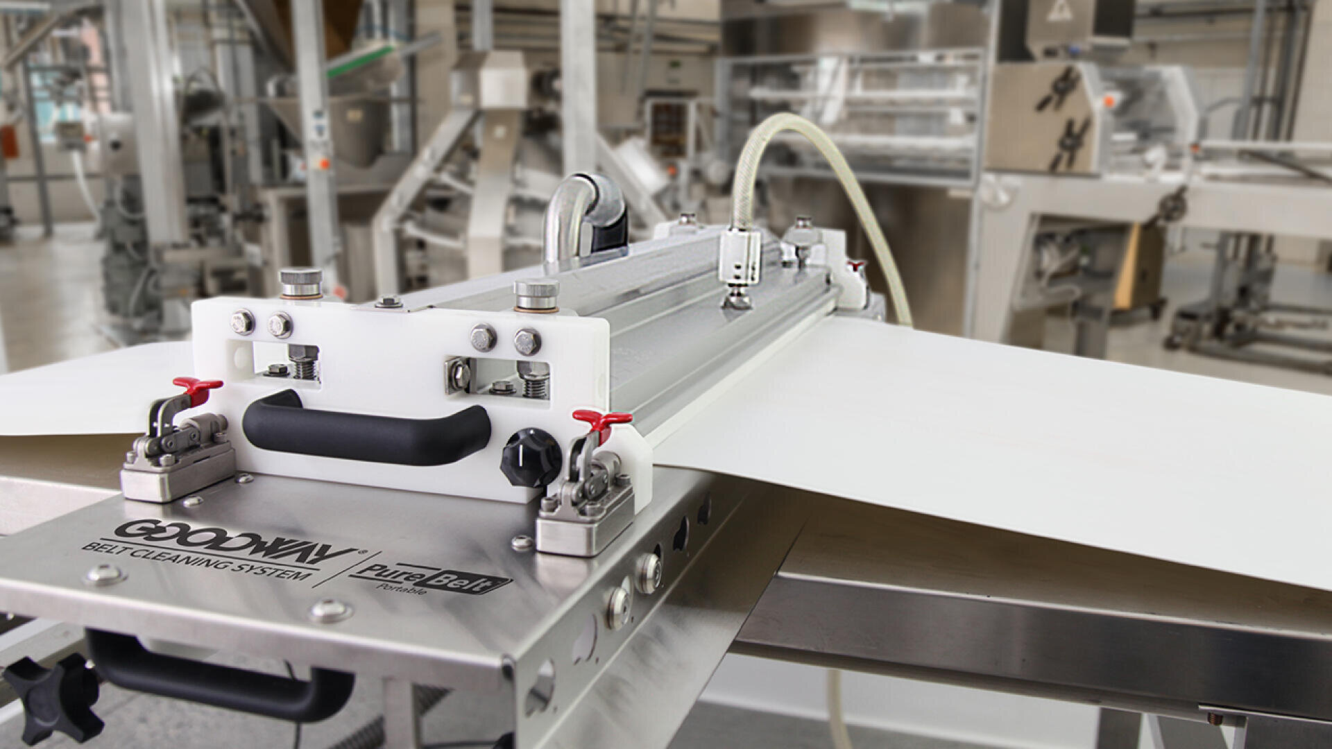 Close-up of an industrial belt cleaning system labeled Goodway PureBelt in a factory setting, with machinery and production equipment visible in the background.