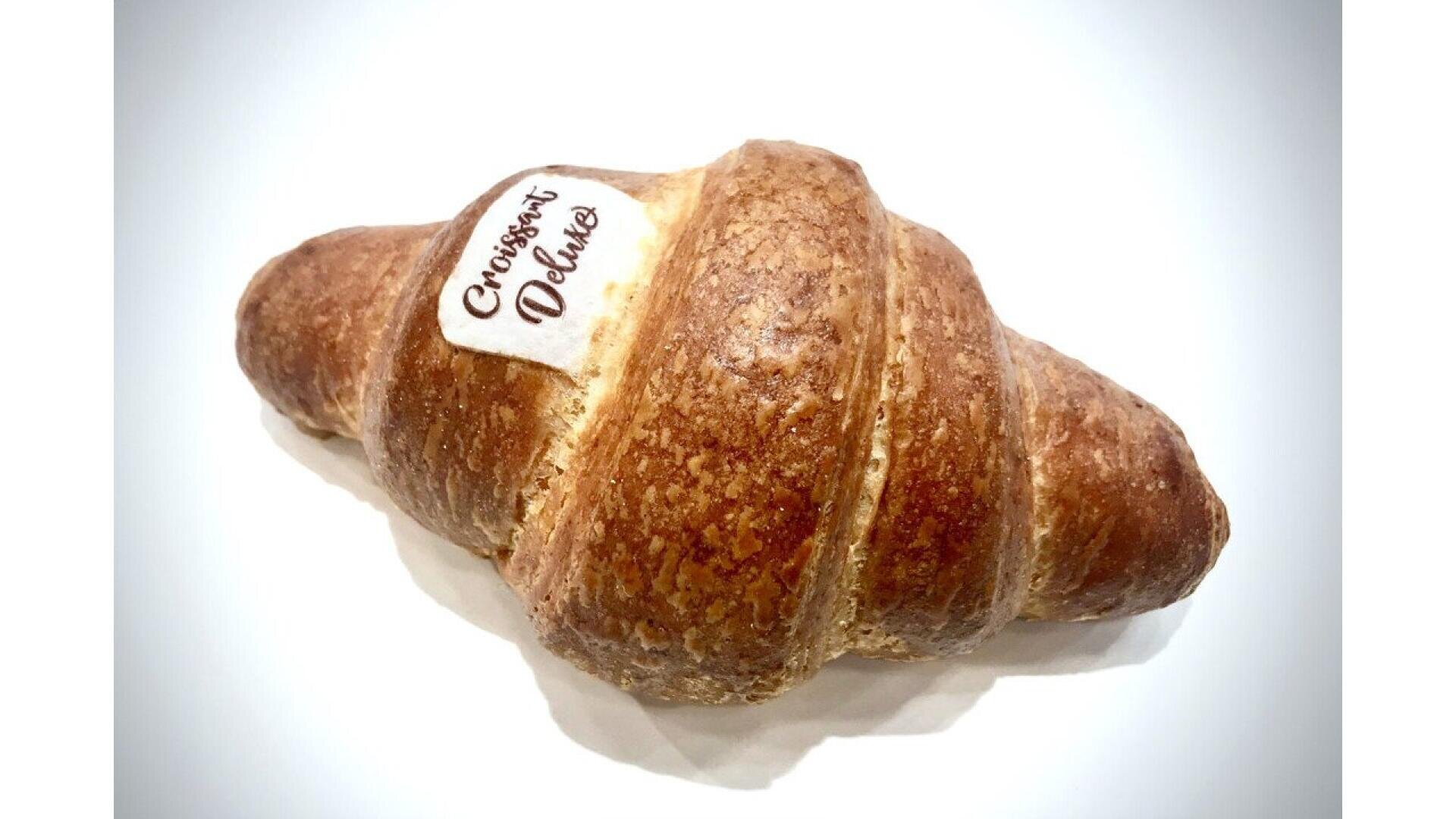 A single croissant with a golden-brown, flaky crust and a white label on top that reads Croissants Deluxe, placed against a white background.