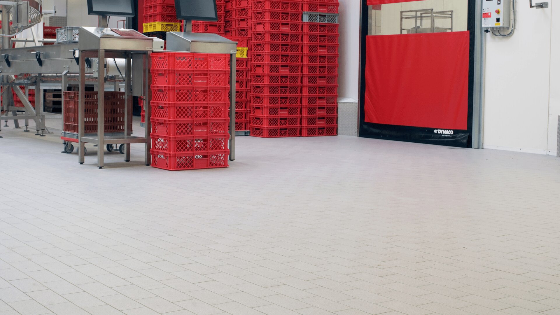 Ein sauberer Industrieraum mit roten Plastikkisten, die neben Tischen und Geräten aus Edelstahl gestapelt sind. Der Boden ist gefliest, und im Hintergrund ist eine große rote Tür zu sehen.