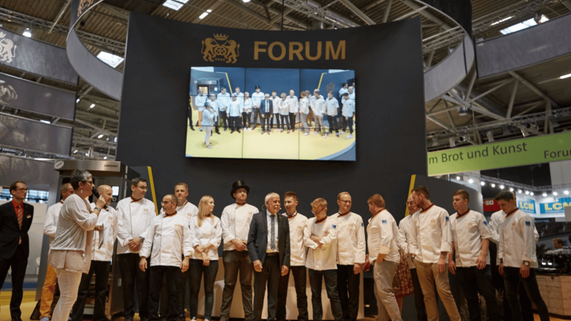 Eine Gruppe von Personen in weißen Kochmänteln steht auf einer Bühne unter einem FORUM-Schild. Über ihnen befindet sich ein großer Bildschirm, der ein weiteres Gruppenfoto zeigt. Die Umgebung scheint eine professionelle Veranstaltung oder Ausstellung zu sein.