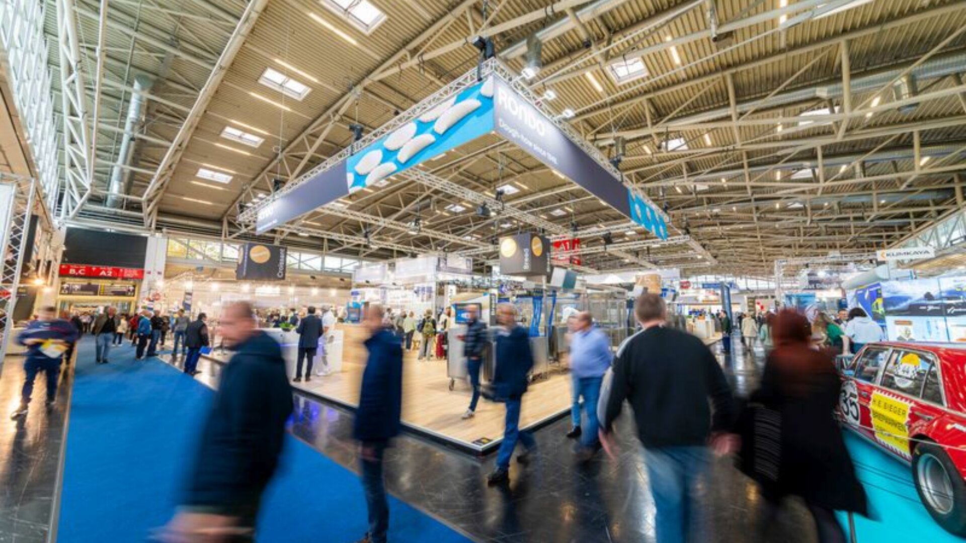 Eine belebte Ausstellungshalle mit Menschen, die unter einer hohen, industriellen Decke mit Oberlichtern an verschiedenen Ständen und Auslagen vorbeilaufen. Auf der rechten Seite ist ein rotes Auto zu sehen, und blaue Teppiche säumen die Gänge.