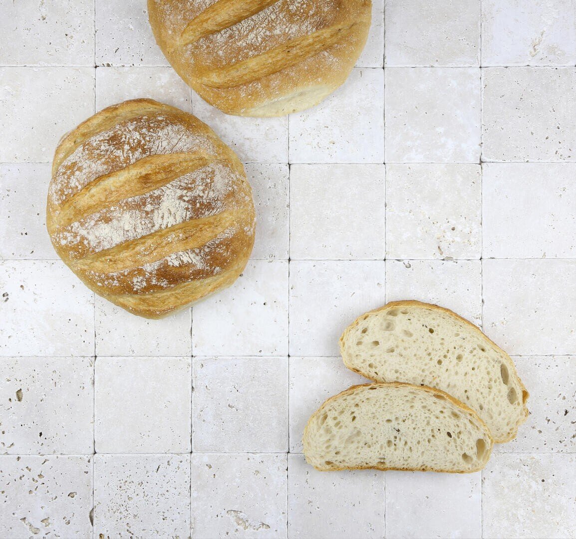 Zwei runde, rustikale Brote und zwei Brotscheiben liegen auf einer hellen, gefliesten Fläche und zeigen eine goldene Kruste und ein luftiges Inneres.