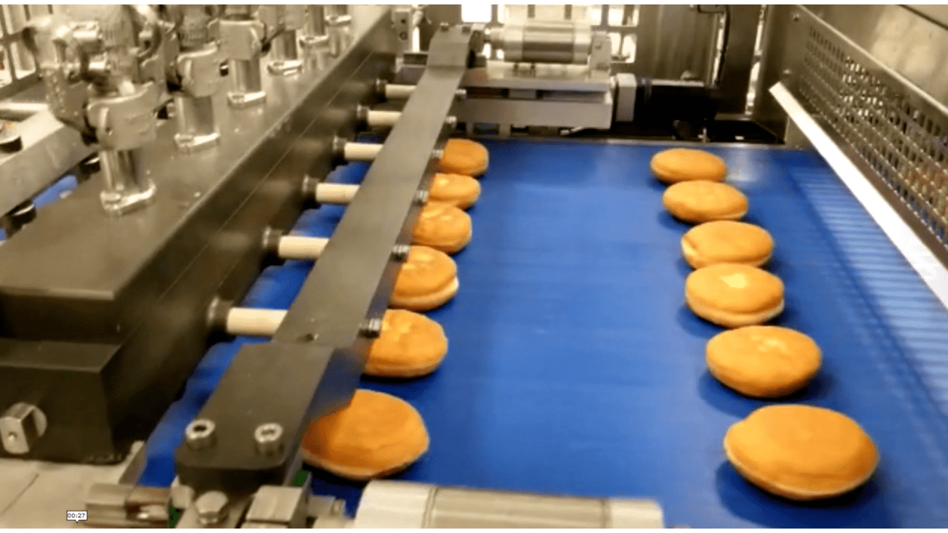 Ein Förderband in einer Fabrik transportiert Reihen von runden, goldenen Sandwichbrötchen unter automatisierten Metallmaschinen, wahrscheinlich zum Abfüllen oder Verpacken.