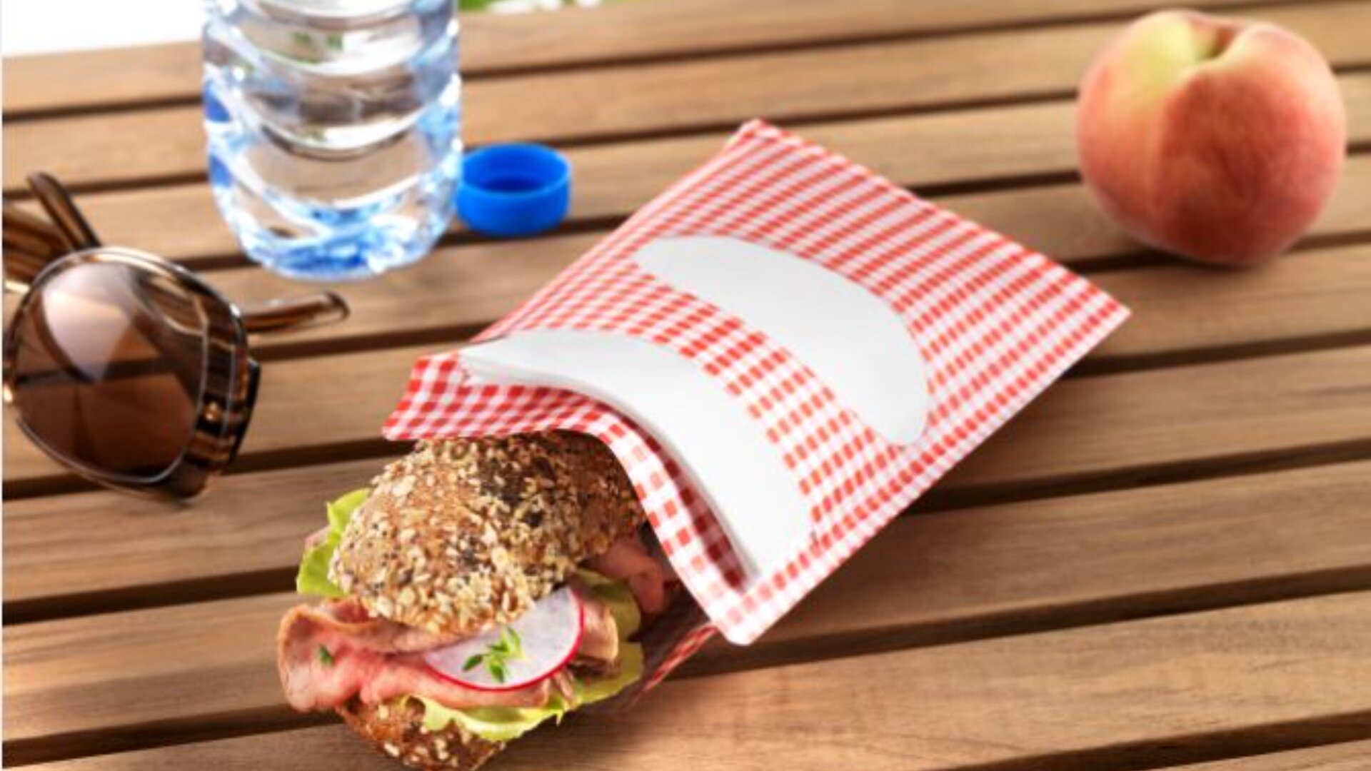 A multigrain sandwich with lettuce, radish, and meat sits partly in a red-and-white checkered wrapper on a wooden table, next to sunglasses, a water bottle, and a peach.