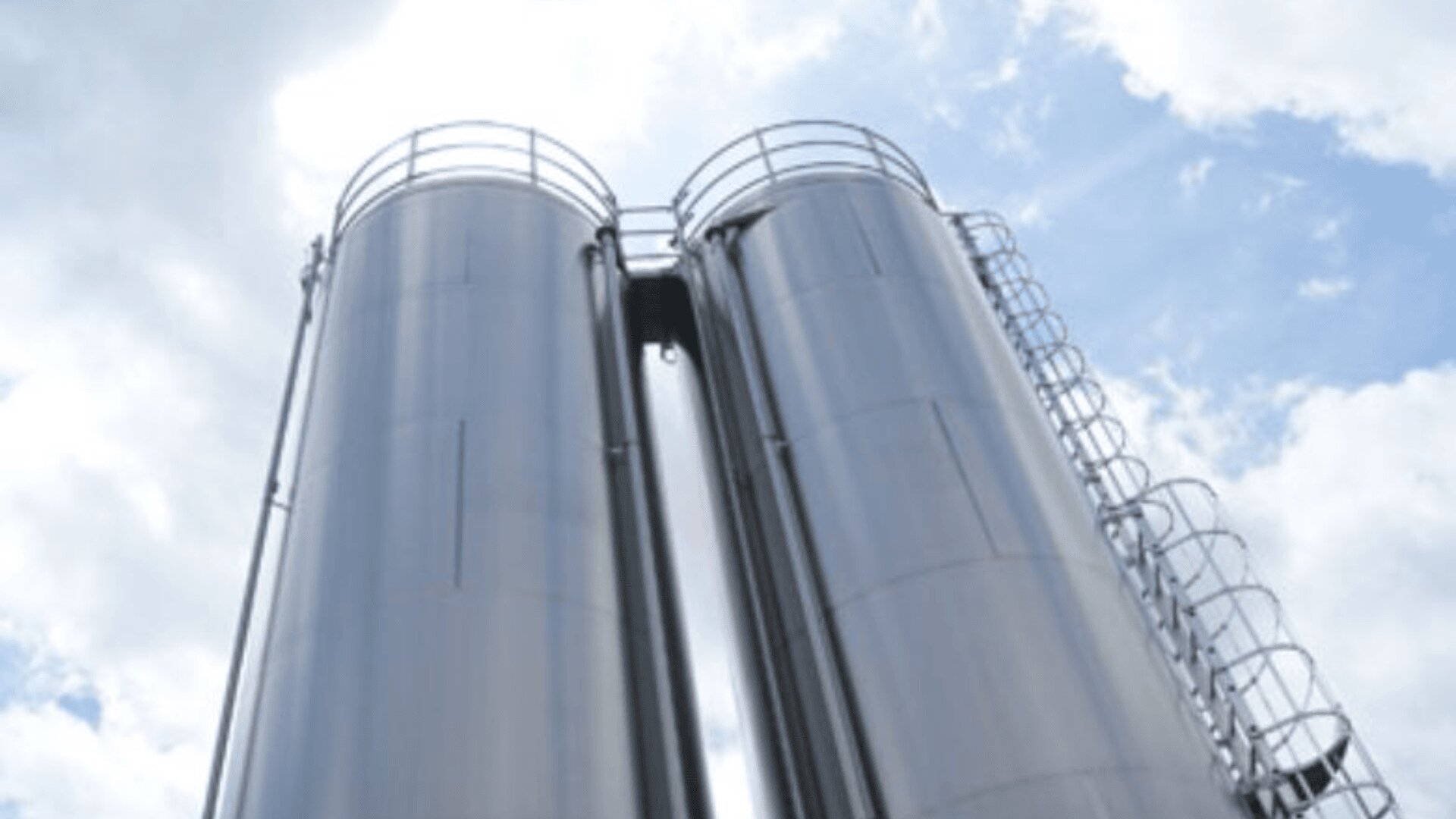 Zwei hohe, zylindrische Metallsilos mit Leitern und Geländern stehen nebeneinander vor einem teilweise bewölkten Himmel.