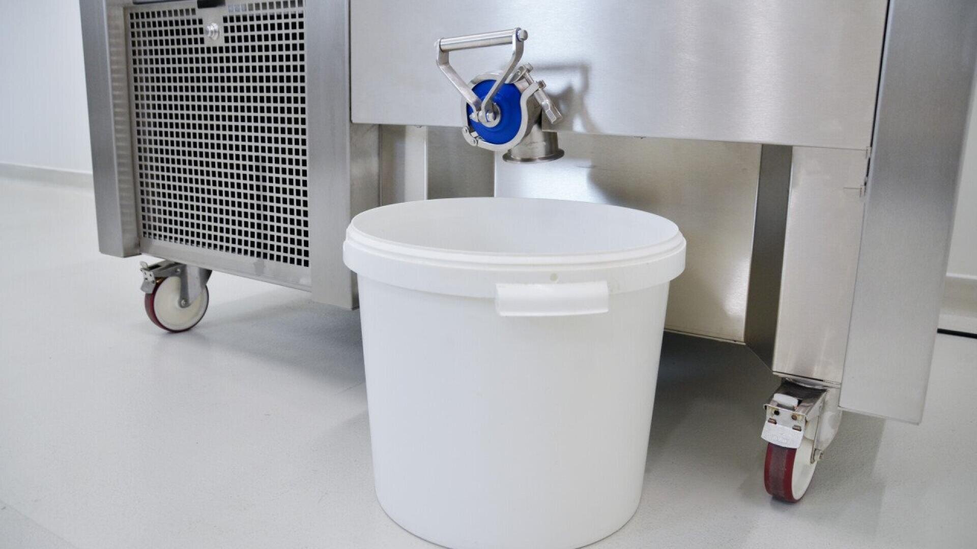 Ein großer weißer Plastikeimer wird unter einen Metallauslauf gestellt, der an einem industriellen Edelstahlgerät auf Rädern befestigt ist, das auf einem sauberen, hellen Boden steht.