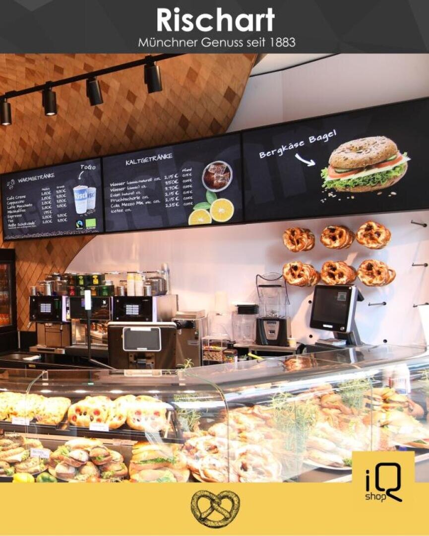 In der Bäckerei Rischart in München werden belegte Brötchen, Gebäck und Brezeln angeboten, über der Theke hängt eine Speisekarte und an der Wand hängen Brezeln. Das Ambiente ist hell und einladend, die Einrichtung modern.