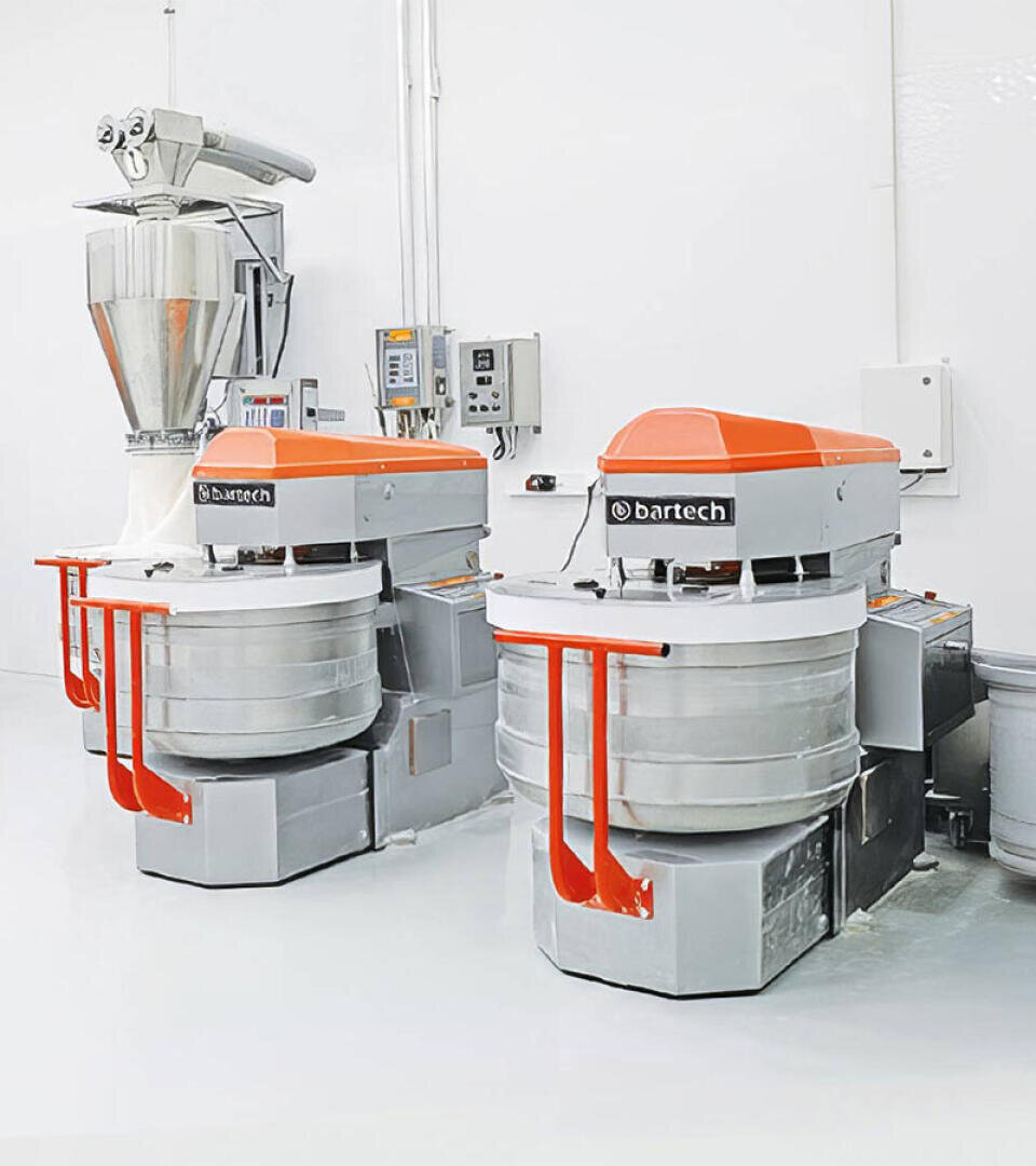 Zwei große industrielle Teigknetmaschinen mit orangefarbenen Deckeln und Schüsseln aus Edelstahl stehen in einem sauberen, weißen Raum. Im Hintergrund sind Schalttafeln und ein Trichter aus Edelstahl zu sehen.