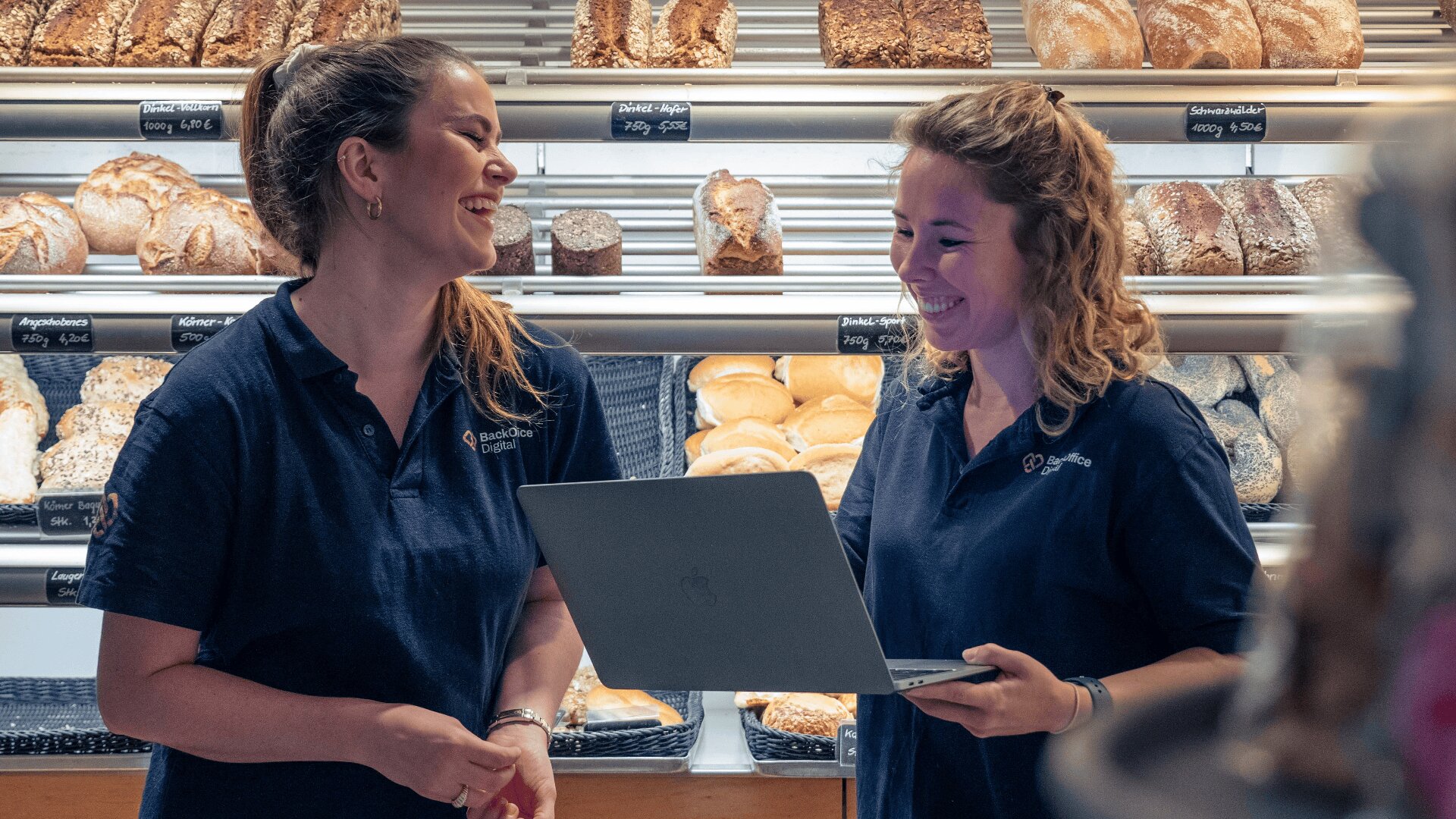 Zwei Frauen in Marineuniform stehen in einer Bäckerei, lächeln und lachen und schauen auf einen Laptop. Die Regale hinter ihnen sind mit Brot und Backwaren gefüllt.