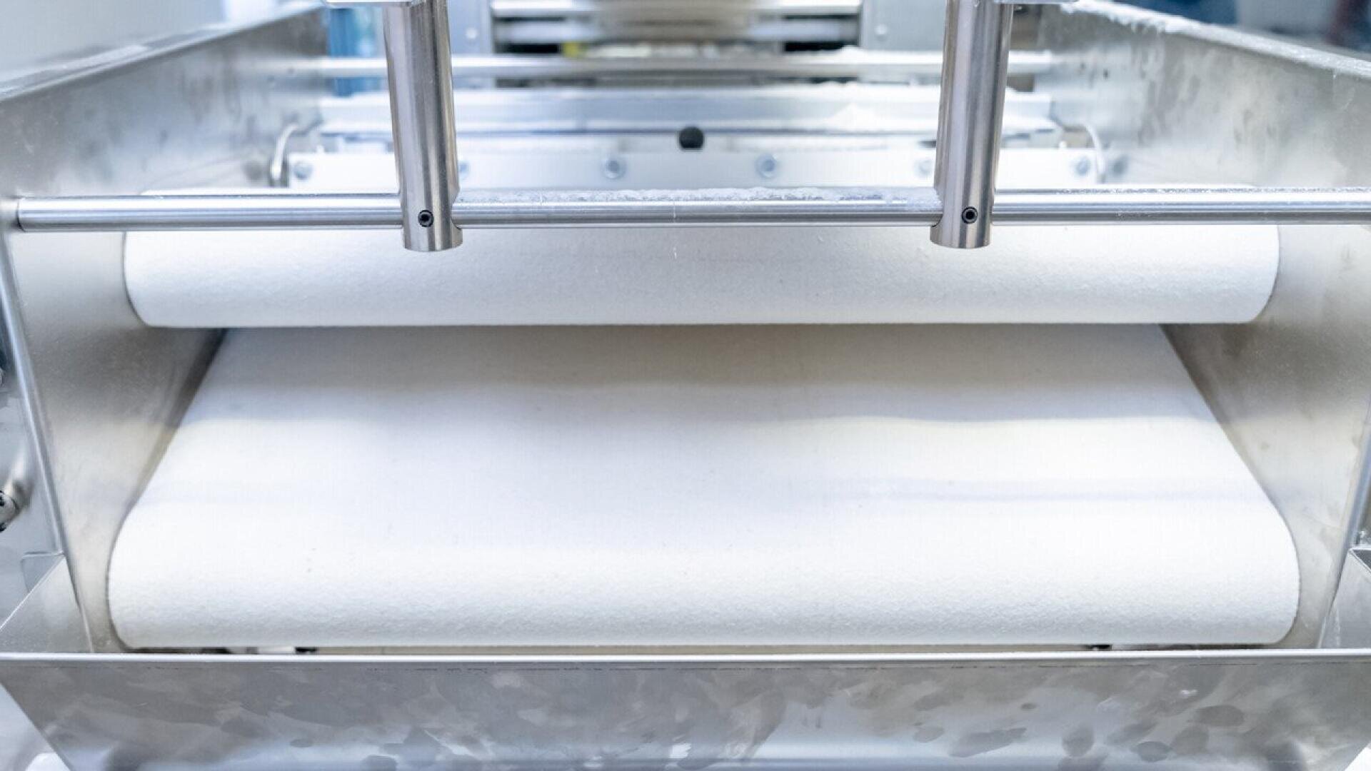 Nahaufnahme einer industriellen Teigausrollmaschine mit zwei Walzen, die den Teig in einer Maschine aus rostfreiem Stahl zu einem dünnen, gleichmäßigen Band pressen und glätten.