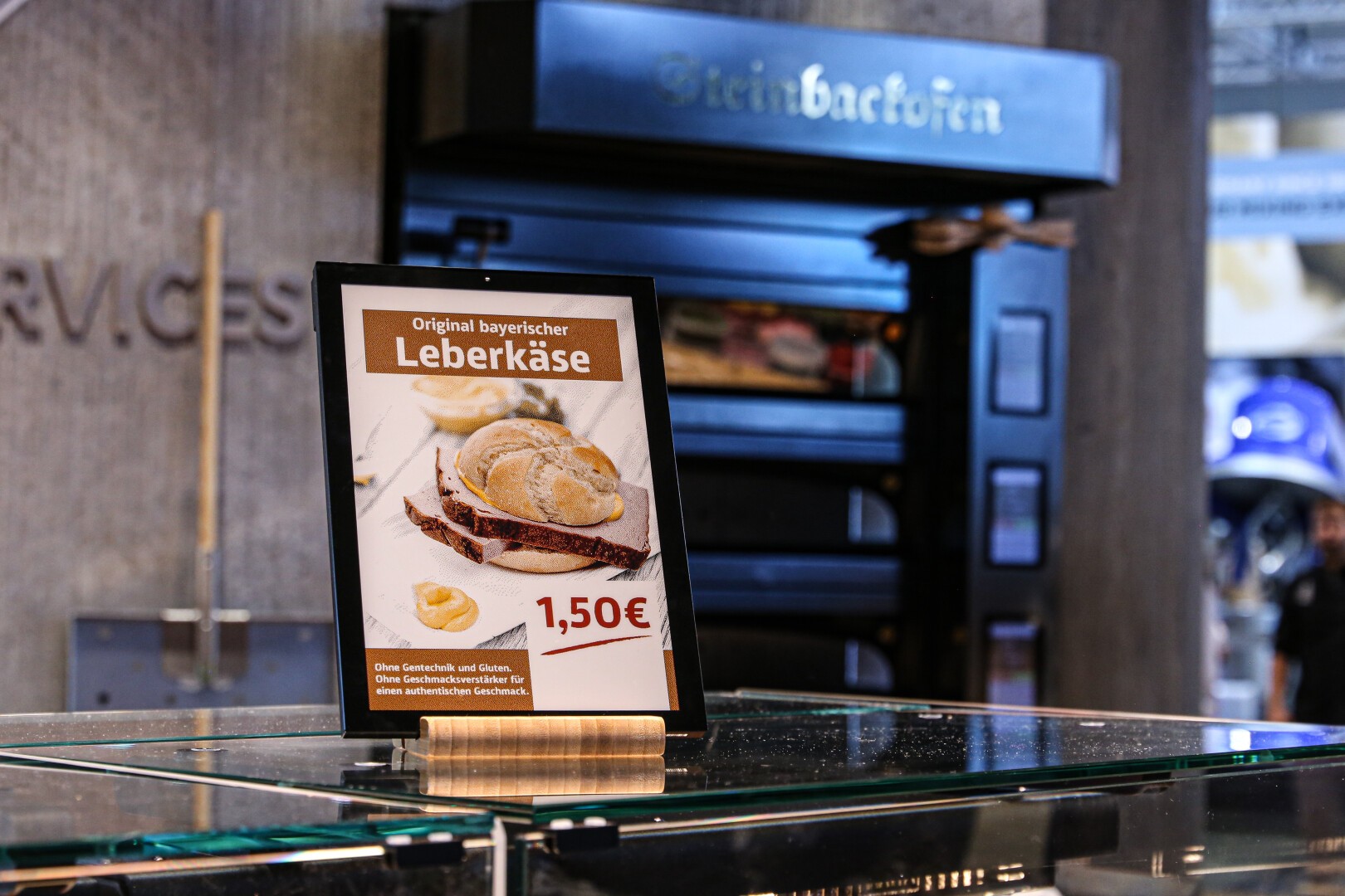 Ein Schild in deutscher Sprache wirbt für Original bayerischen Leberkäse für 1,50 Euro. Es zeigt ein Sandwich mit Leberkäsescheiben und Senf. Das Schild steht auf einer Glastheke vor einem dunklen Backofen.