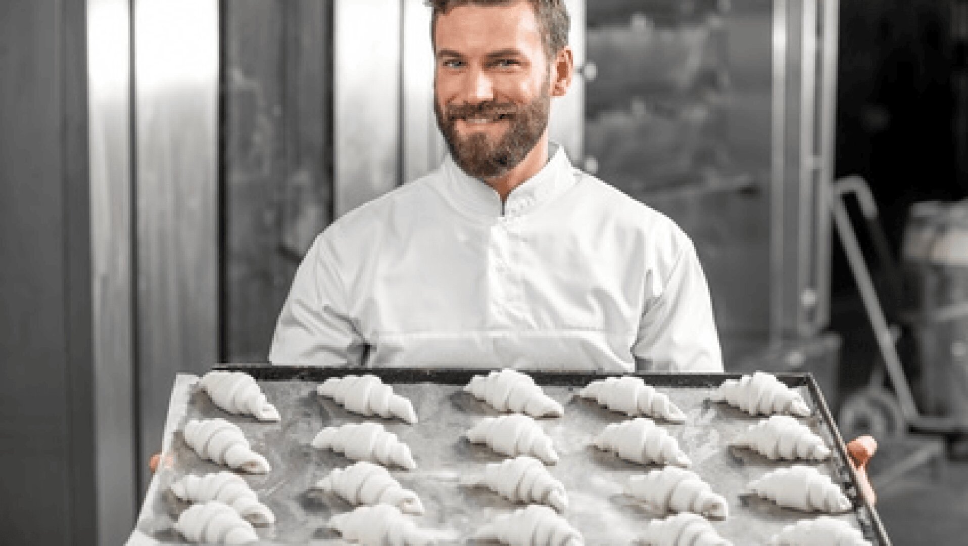 Ein lächelnder Bäcker im weißen Kochkittel hält ein großes Tablett mit ungebackenen Croissants in einer Großküche.