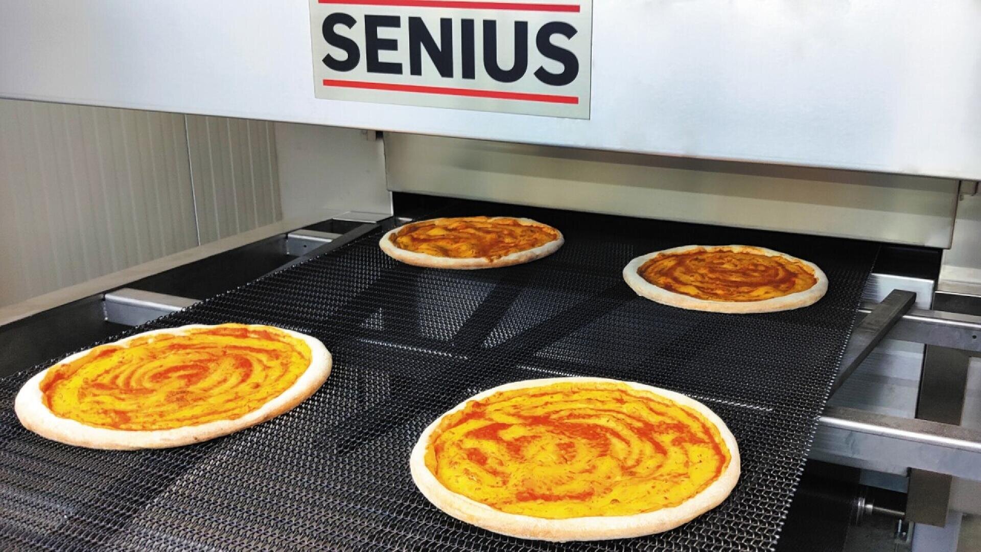 Four uncooked pizza bases with tomato sauce are moving on a conveyor belt into a machine labeled SENIUS, likely in a commercial pizza production facility.