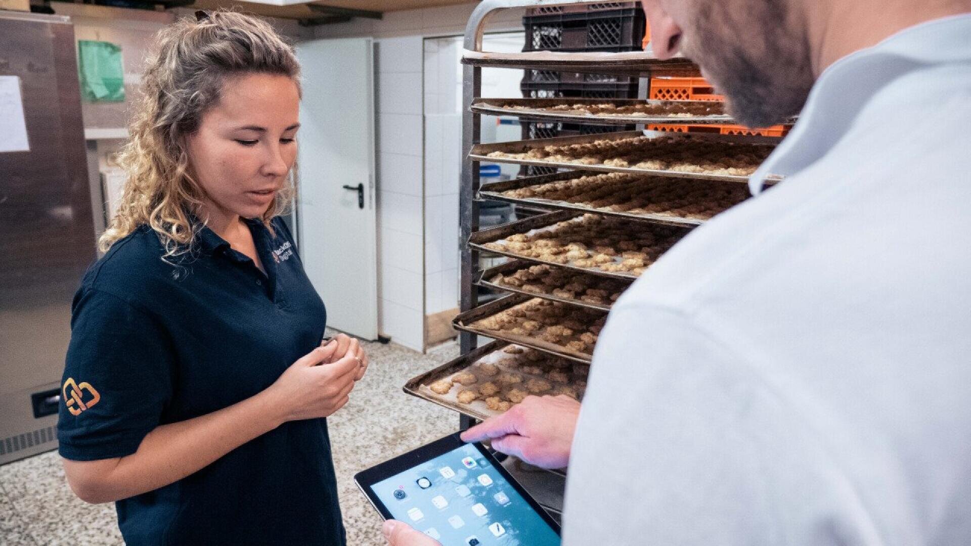 A woman stands near baking trays filled with cookies while a man uses a tablet. They appear to be discussing or inspecting the baked goods in a commercial kitchen setting.