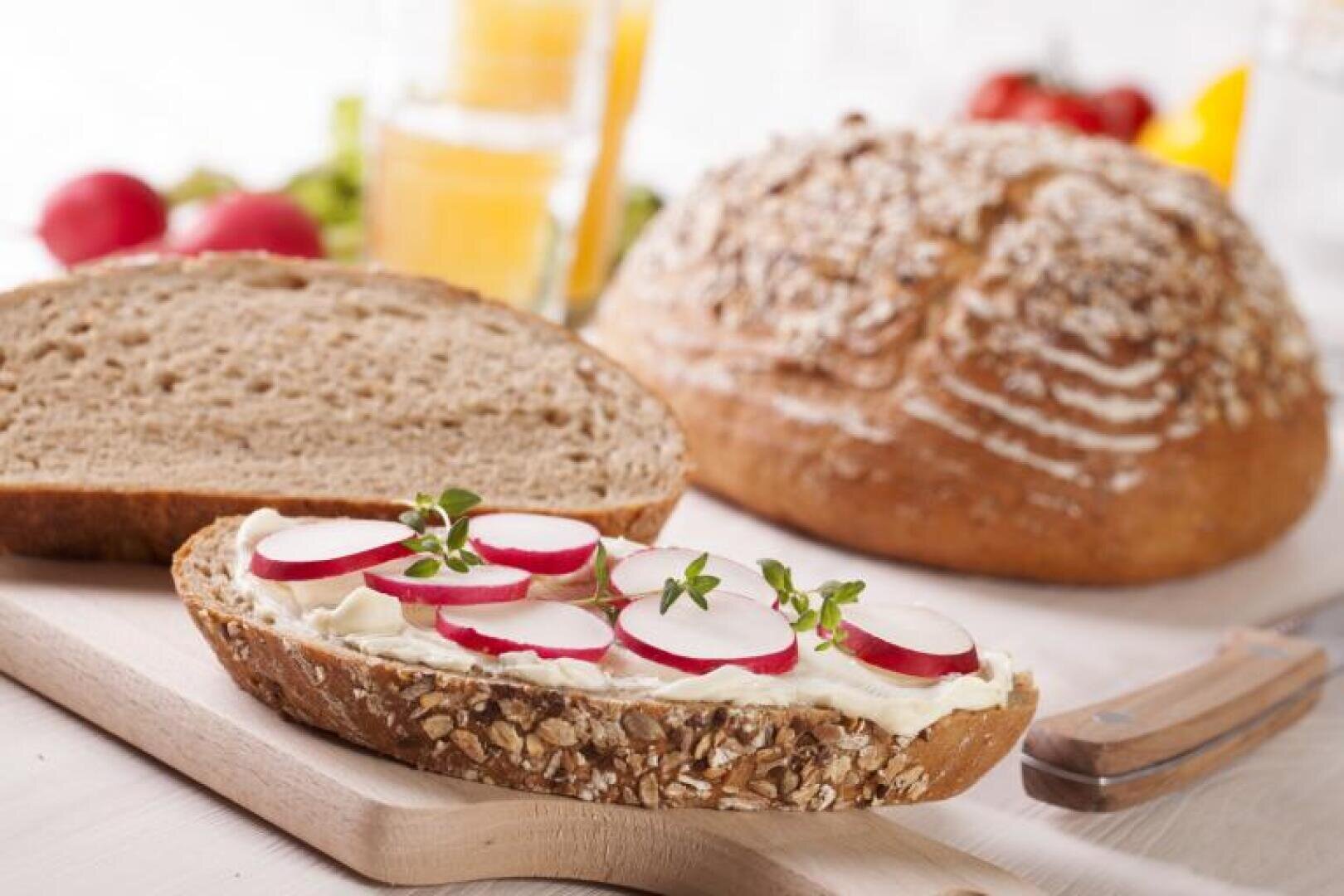 A slice of whole-grain bread with cream cheese, radish slices, and fresh herbs lies on a cutting board, next to a loaf of bread and a glass of orange juice.