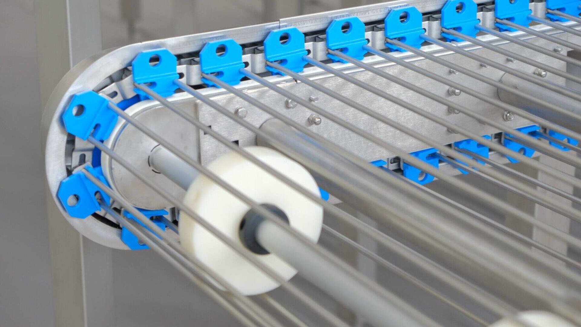 Nahaufnahme einer Industriemaschine aus rostfreiem Stahl mit parallelen Metallstäben, blauen Kunststoffklammern und weißen kreisförmigen Walzen, wahrscheinlich Teil einer automatisierten Verarbeitungs- oder Fertigungsanlage.