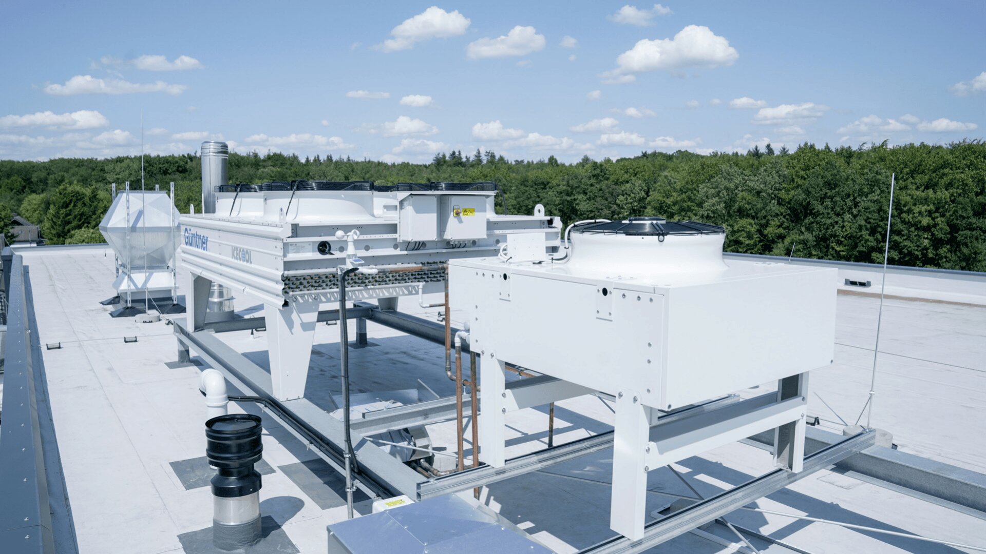 Large industrial HVAC units are installed on a flat rooftop under a clear blue sky with scattered clouds, surrounded by trees in the background.
