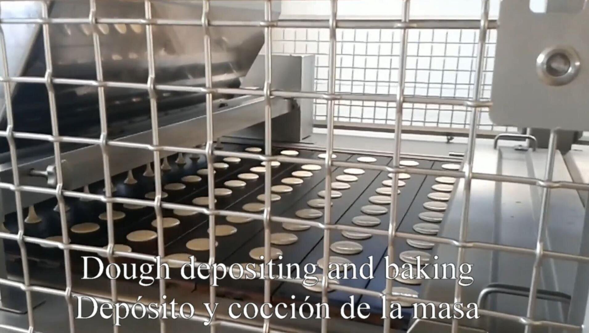 A metal machine deposits rows of round dough pieces onto a conveyor belt, seen through a metal safety grid; English and Spanish subtitles read “Dough depositing and baking / Depósito y cocción de la masa.”.