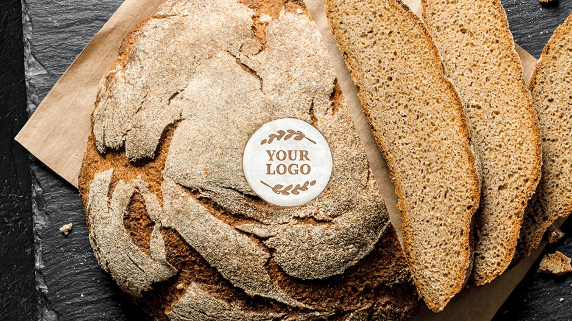 Ein runder, rustikaler Brotlaib mit einem Papierkreis, der mit IHREM LOGO beschriftet ist, neben mehreren Scheiben desselben Brotes, die auf braunem Pergamentpapier und einer dunklen Oberfläche liegen.
