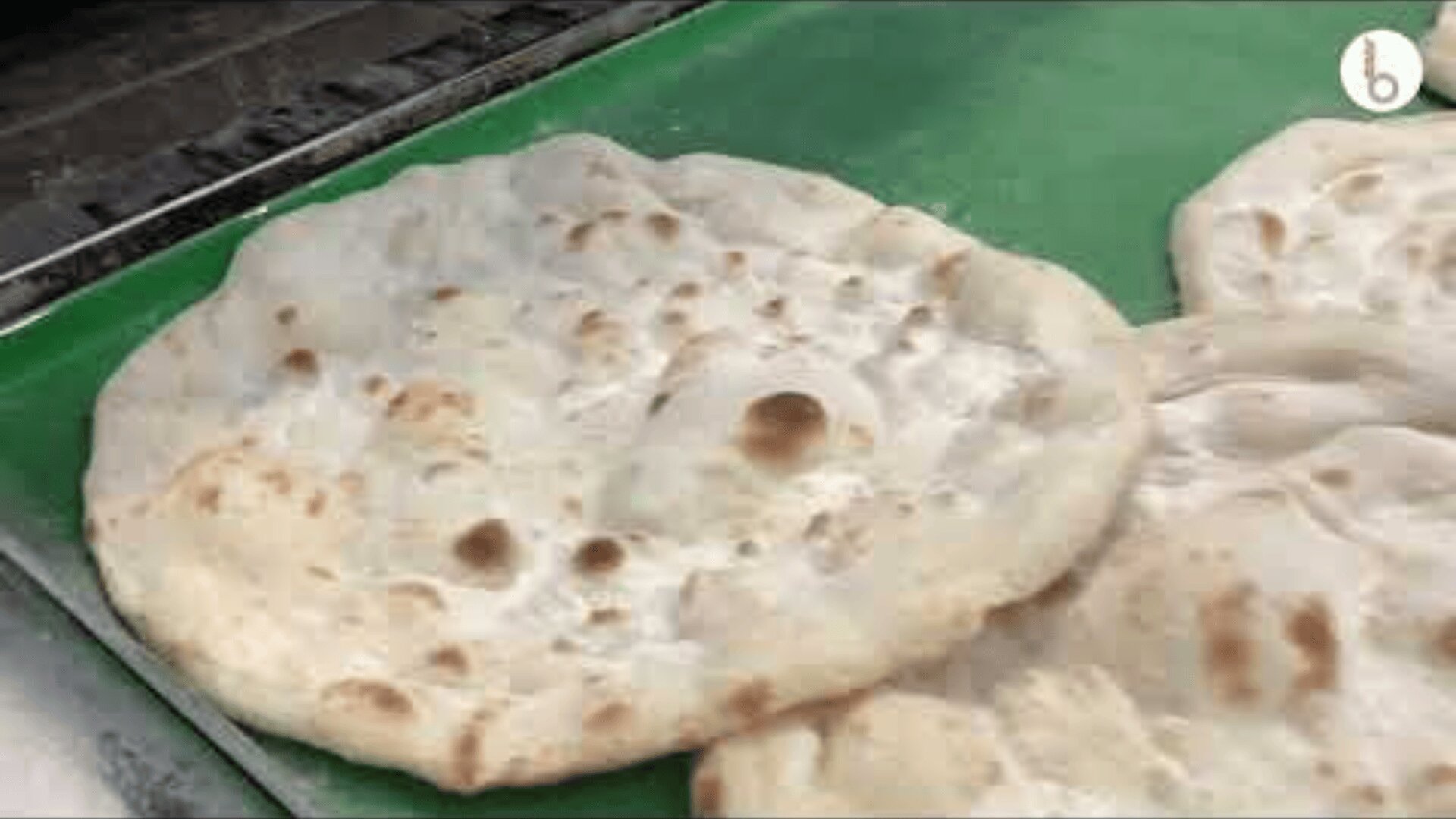 Fladenbrote mit goldbraunen Flecken werden auf einer grünen Fläche neben einem Grill platziert. Die Brote wirken frisch gebacken und sind an manchen Stellen leicht aufgeblasen.