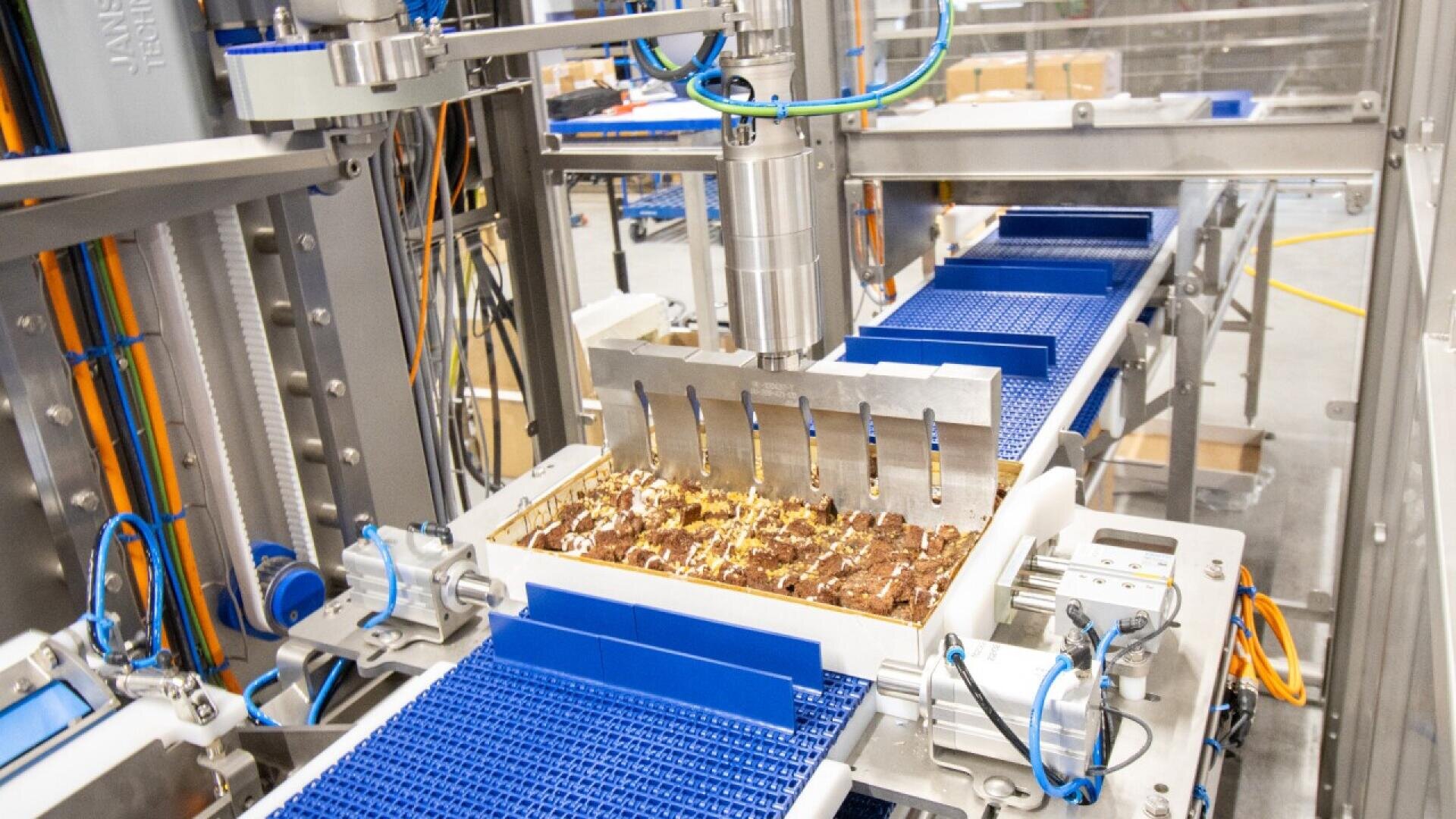 Eine Fabrikmaschine mit blauen Förderbändern verarbeitet und ordnet Reihen von Schokoladen- oder Schokoriegeln unter einem Roboterarm in einer industriellen Umgebung an.
