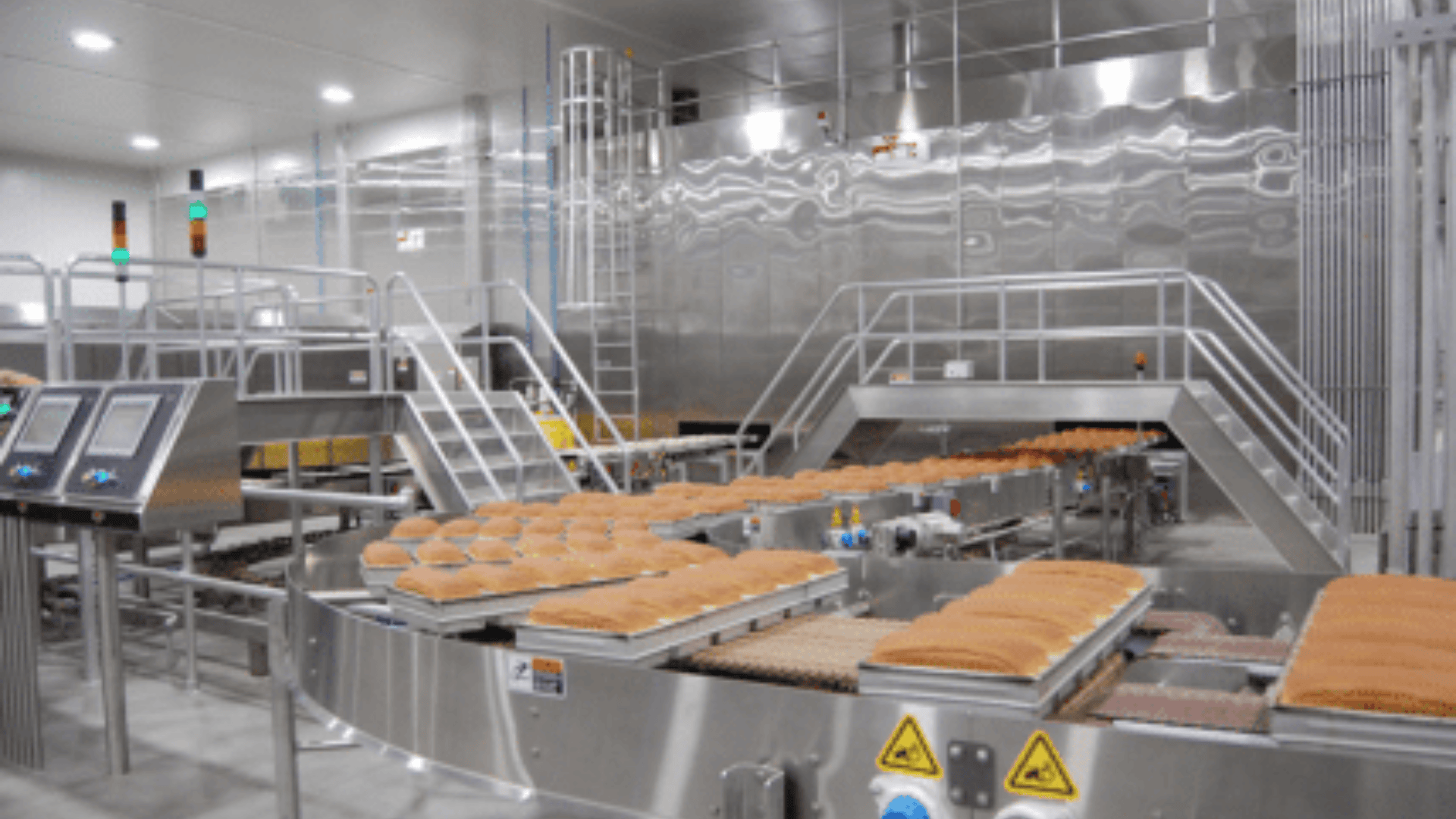 Eine moderne Industriebäckerei aus rostfreiem Stahl mit Förderbändern, auf denen reihenweise verpackte Brotlaibe transportiert werden, Schalttafeln, Sicherheitsschildern und einer sauberen, gut beleuchteten Umgebung.