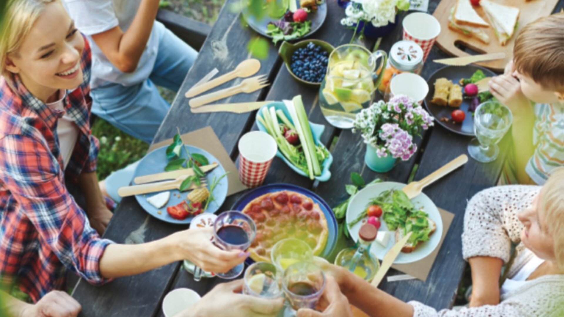 A group of people sit around an outdoor table, sharing a meal with fresh fruit, salad, and drinks. They are toasting, smiling, and enjoying a casual, colorful picnic with flowers and tableware.