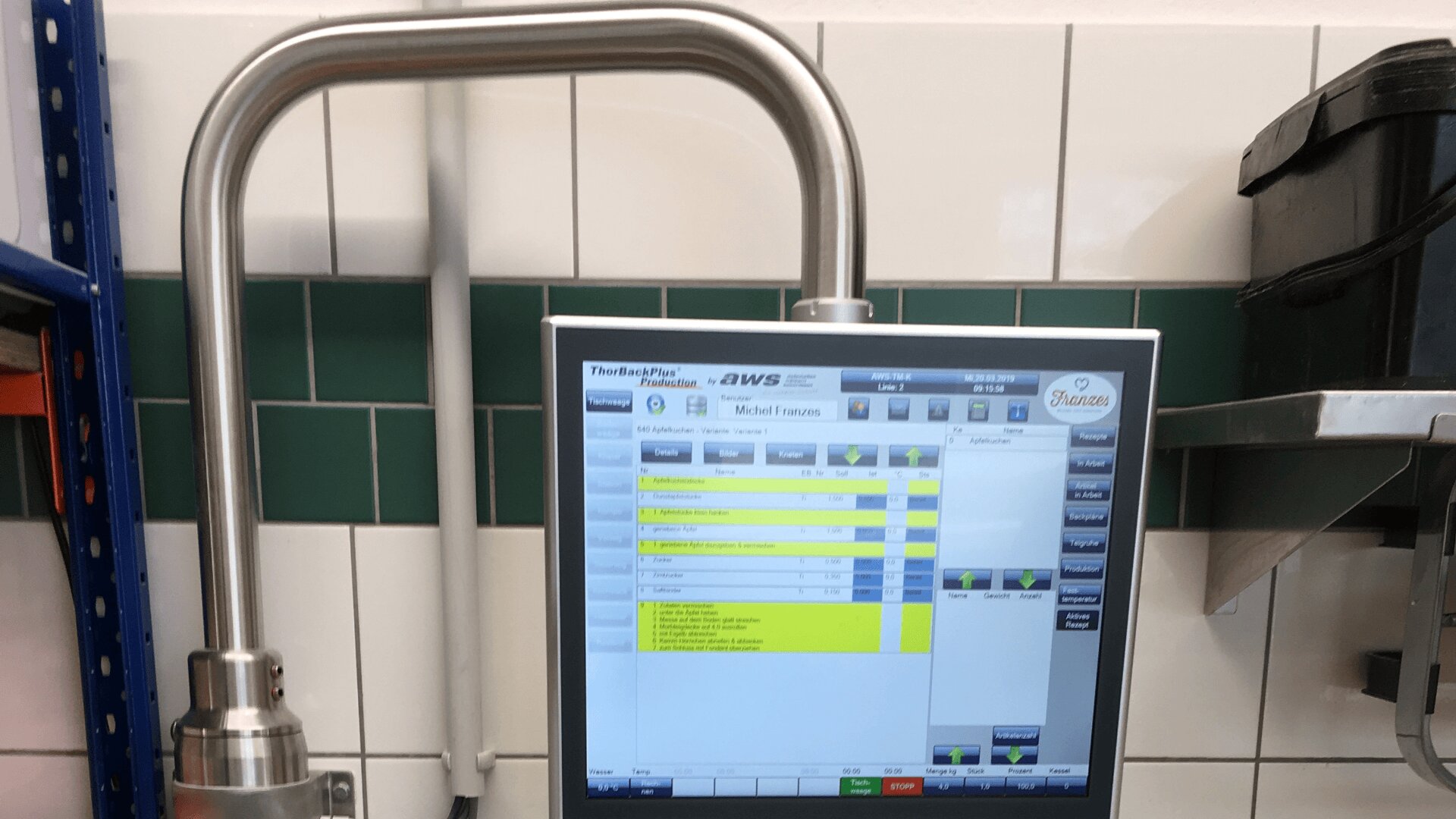 Ein Touchscreen-Bedienfeld mit einem Metallrahmen zeigt hervorgehobene Menüoptionen an. Der Hintergrund besteht aus weiß und grün gefliesten Wänden, neben denen sich Regale und verschiedene Geräte befinden.
