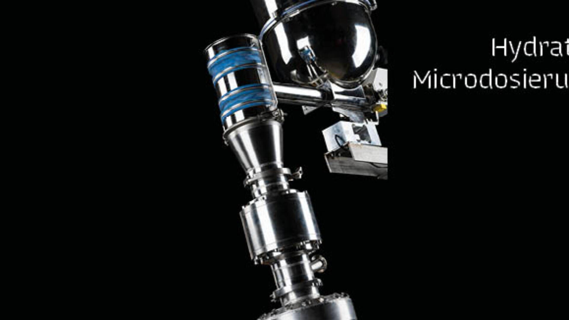Nahaufnahme einer Industriemaschine mit Metallrohren und blauen Komponenten vor einem schwarzen Hintergrund. Ein Teil des weißen Textes lautet Hydra Microdosieru.