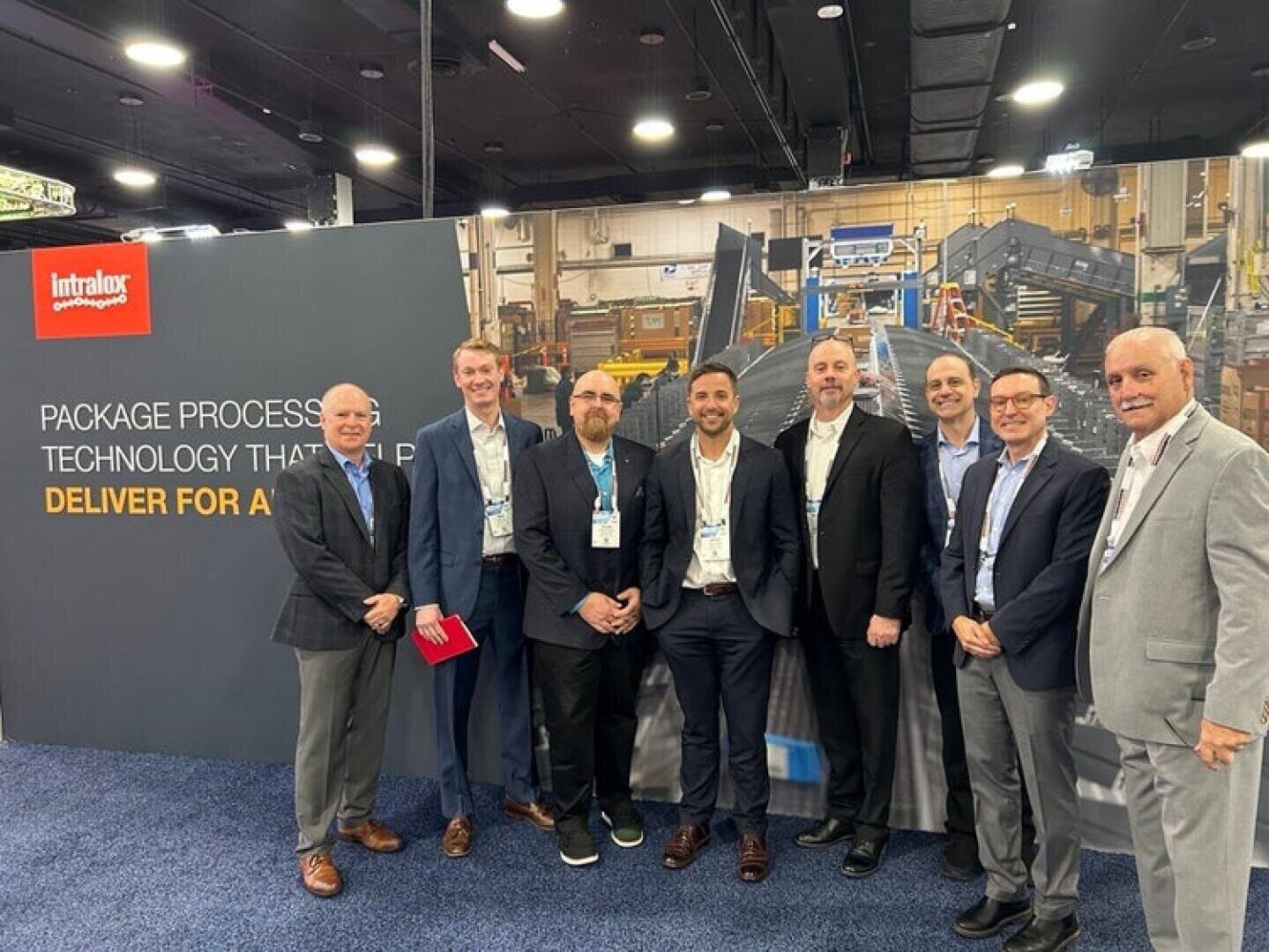 Eight men in business attire stand smiling in front of an Intralox trade show booth with a large display about package processing technology and equipment in a convention center.