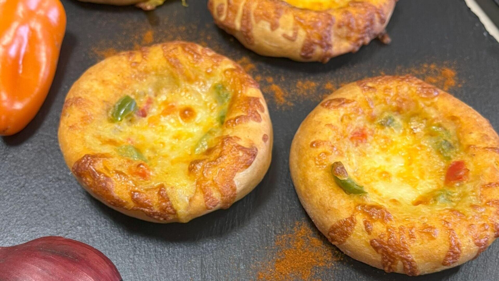 Three small, round, baked bread bowls topped with melted cheese and bits of colorful bell peppers are arranged on a black slate surface next to part of a red onion and an orange pepper.
