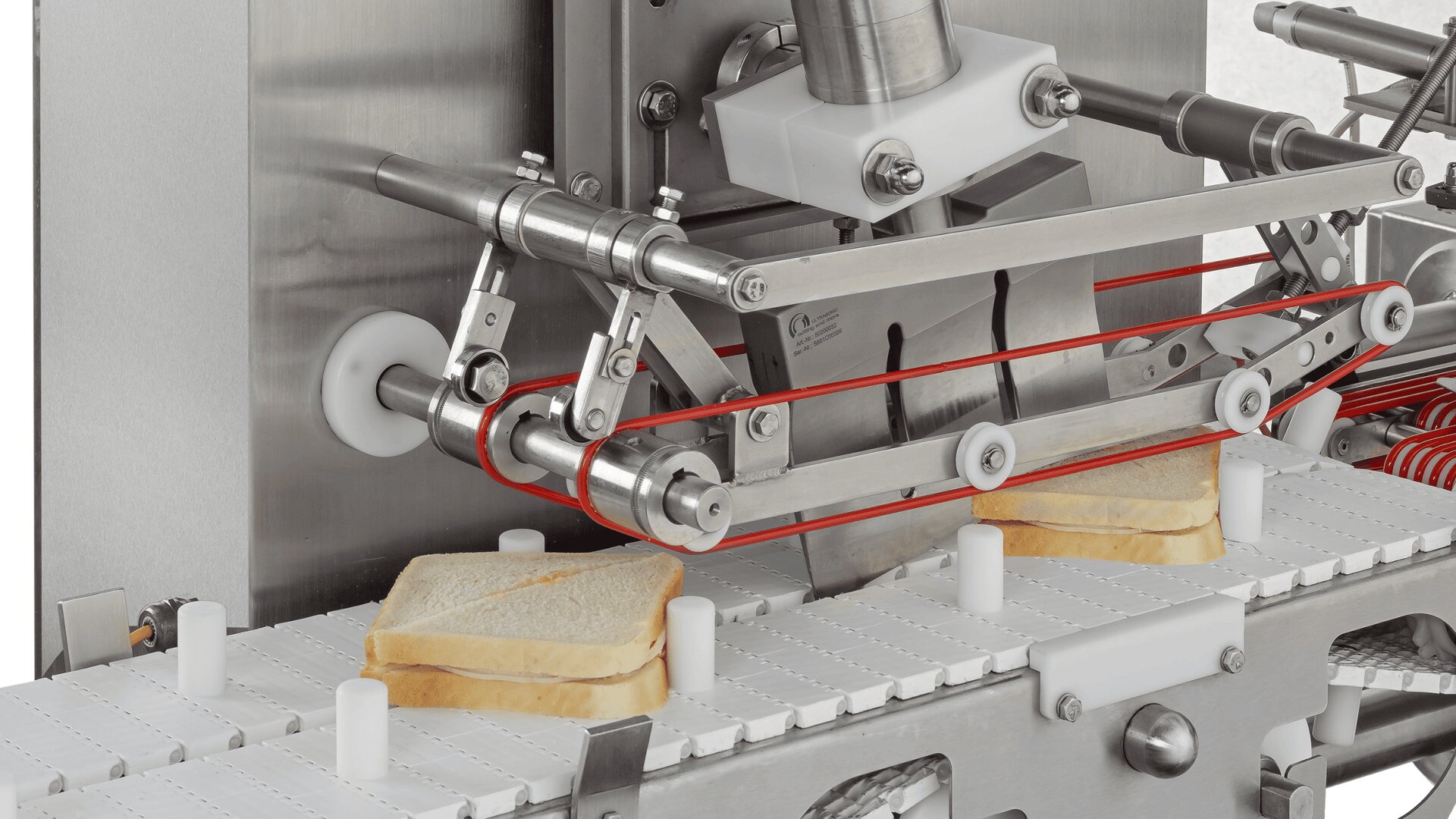 Nahaufnahme einer Lebensmittelverarbeitungsmaschine, die Sandwiches auf einem Förderband schneidet. Zwei Sandwiches mit sichtbarem Brot werden unter einem Schneidemechanismus mit roten Bändern und Metallarmen positioniert.