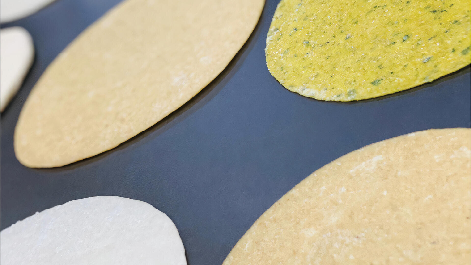 Several round, flat tortillas in different colors—white, yellow, and green—are arranged on a dark surface, viewed close up and at an angle.