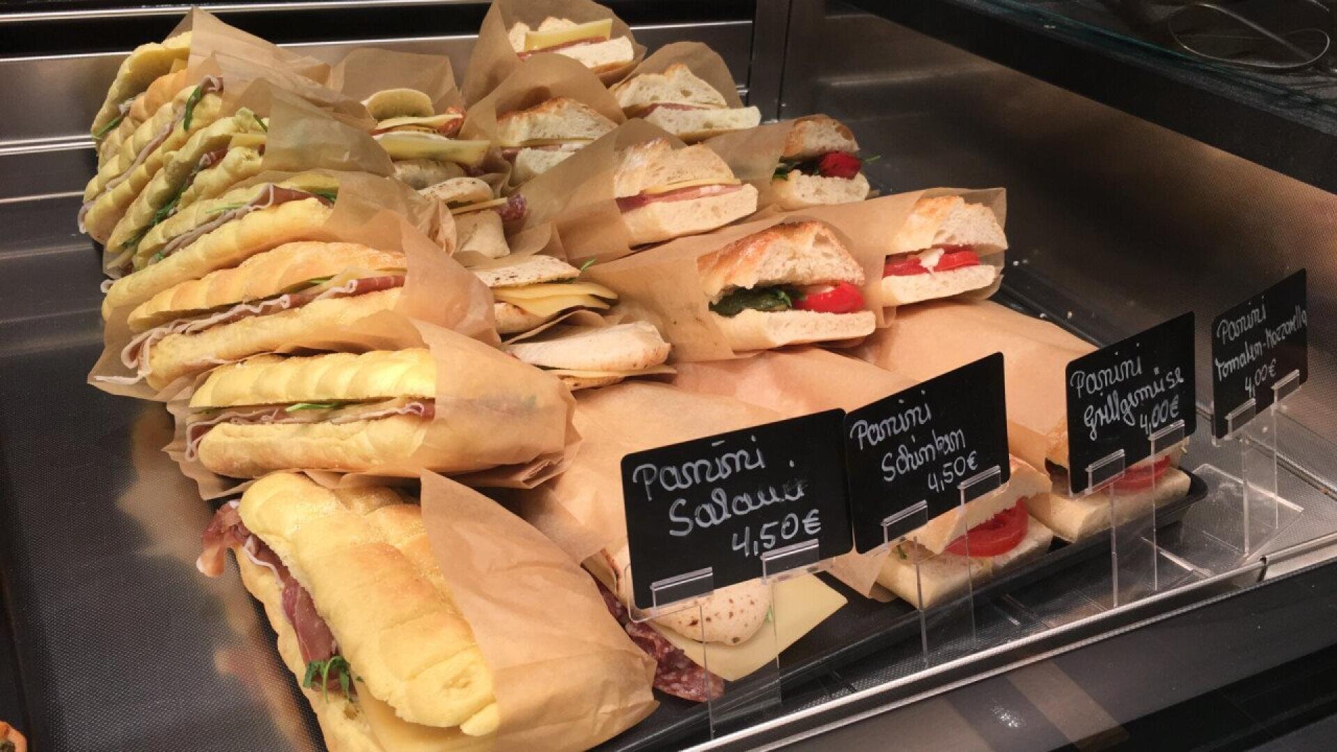 Eine Vitrine mit verschiedenen in Papier eingewickelten Panini-Sandwiches, die mit kleinen Kreideschildern mit Namen und Preisen in Euro gekennzeichnet sind. Die Sandwiches enthalten Zutaten wie Käse, Fleisch, Spinat und Tomaten.