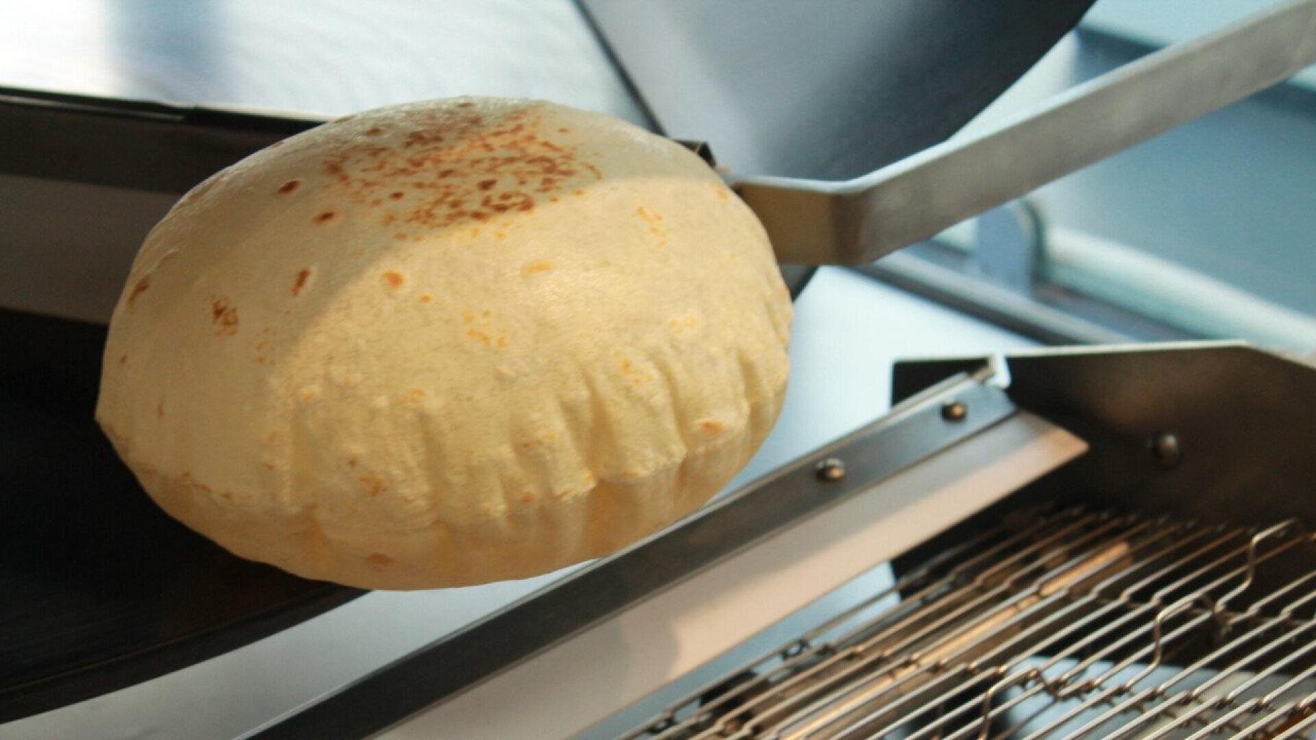 Ein frisch gebackenes, rundes, aufgeblasenes Brot wird mit einem Metallspatel über ein Kühlregal in einer Küche oder Bäckerei gehalten. Das Brot ist luftig und an der Oberseite leicht gebräunt.