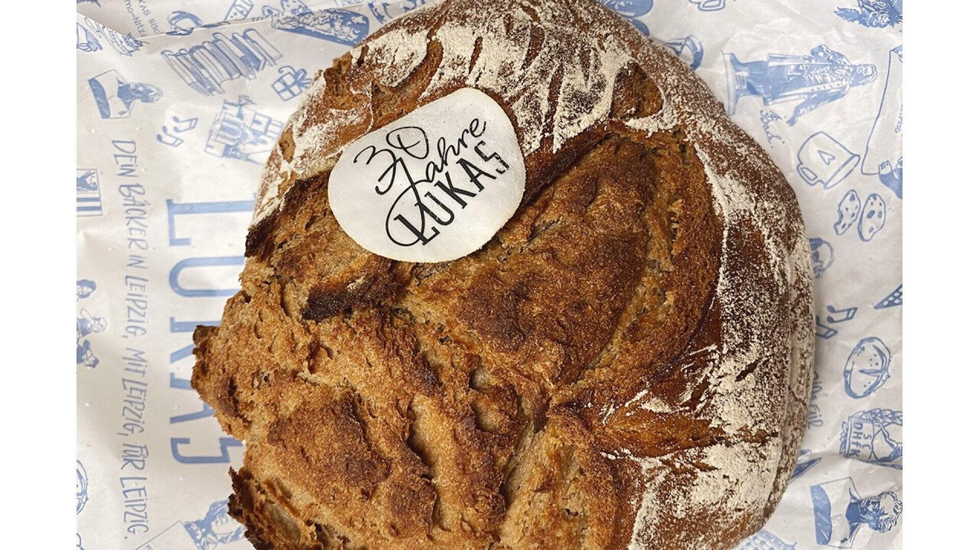 A round loaf of rustic bread with a white sticker that reads “30 Jahre Lukas” placed on top, resting on decorative bakery paper with blue designs and German text.