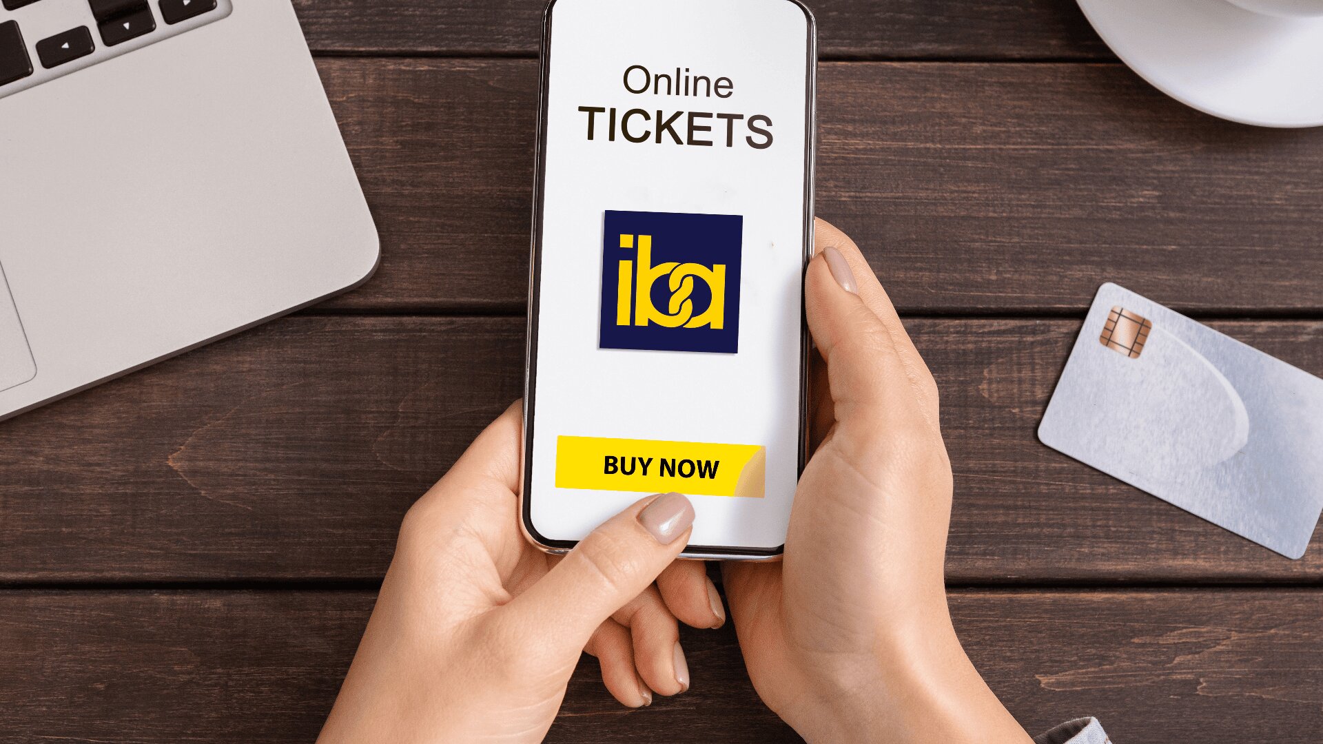 A person holds a smartphone displaying an online ticketing screen with the iba logo and a yellow Buy Now button. A laptop, a cup, and a credit card are on a wooden table in the background.