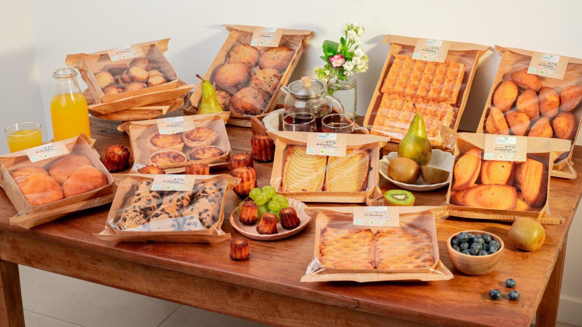 Auf einem Holztisch liegen verpacktes Gebäck, Kuchen und Brot sowie Obst wie Trauben, Birnen, Kiwi und Blaubeeren. Eine Karaffe und ein Glas Orangensaft sind ebenfalls vorhanden, und im Hintergrund stehen kleine Vasen mit Blumen.
