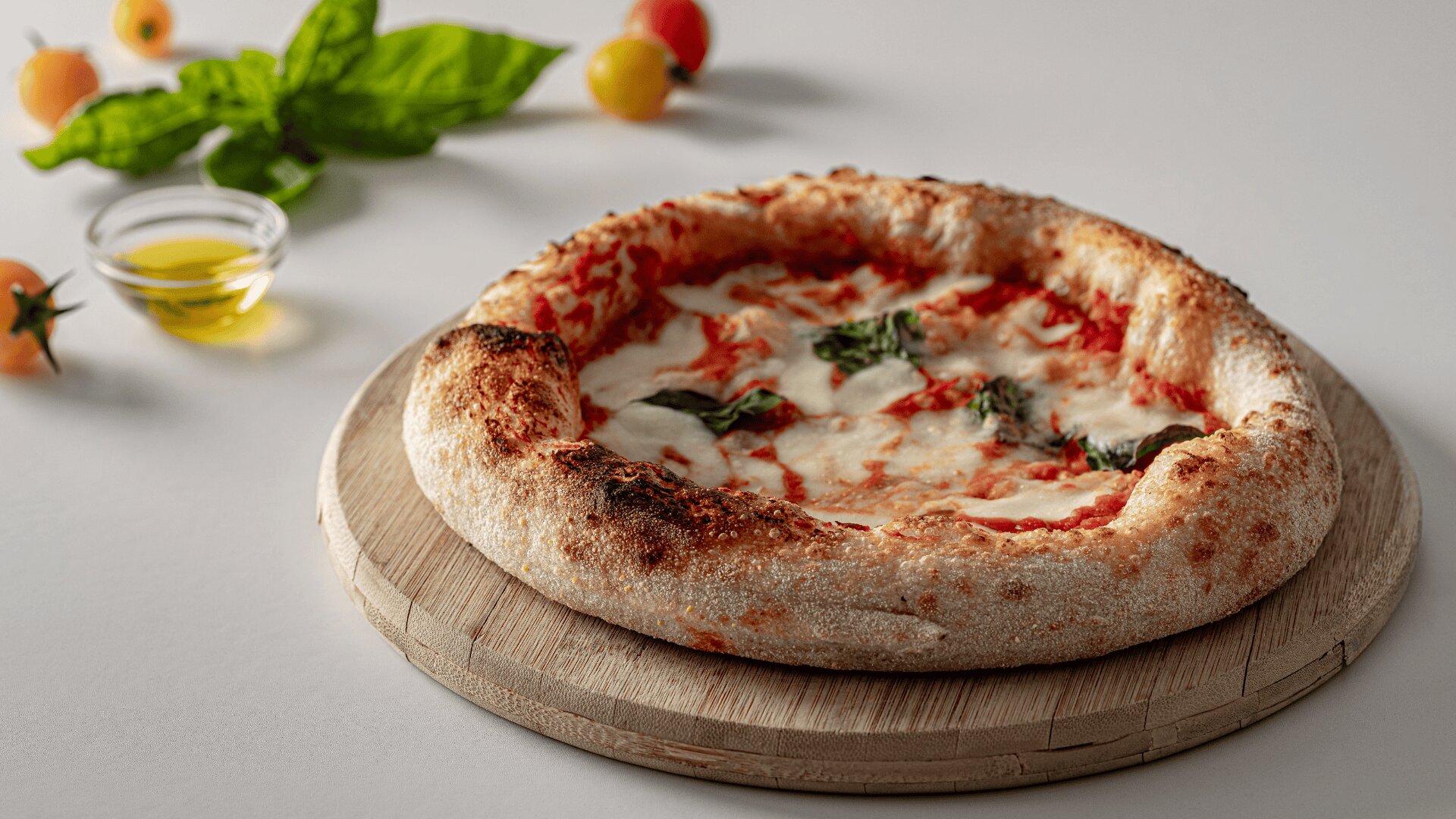 Eine frisch gebackene Margherita-Pizza mit geschmolzenem Käse und Basilikum liegt auf einem Holzbrett. Kirschtomaten, Basilikumblätter und eine kleine Schale mit Olivenöl stehen im Hintergrund auf einer weißen Fläche.