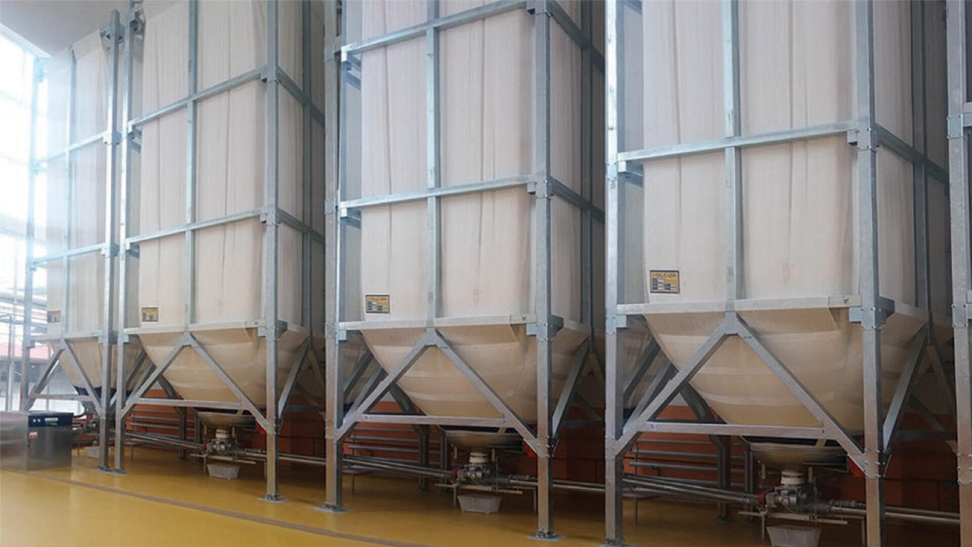 Drei große industrielle Stoffsilos mit Metallrahmen stehen nebeneinander in einer Anlage mit gelbem Boden, die zur Lagerung und Abgabe von Schüttgütern verwendet wird.