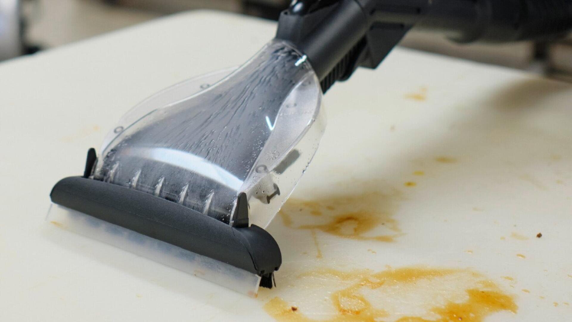 A handheld carpet cleaner is being used on a light-colored stained surface, visibly removing dirt and liquid stains as it cleans.
