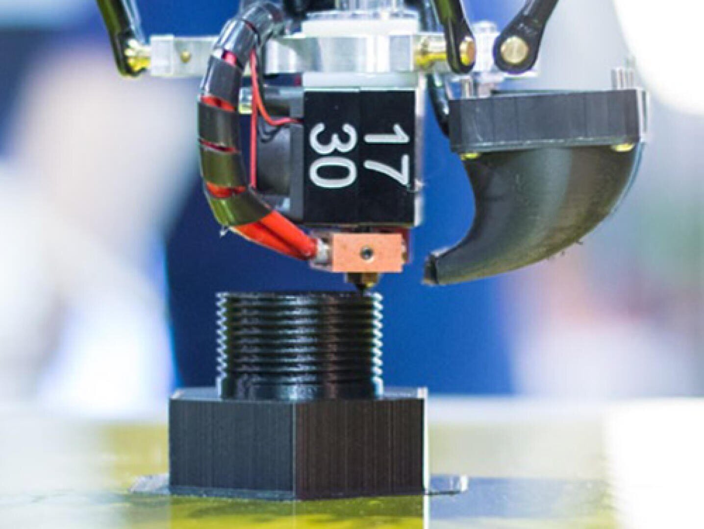 Eine Nahaufnahme einer 3D-Druckerdüse, die schwarzes Filament extrudiert, um ein zylindrisches Objekt mit Gewinde auf der Bauplattform zu erzeugen.