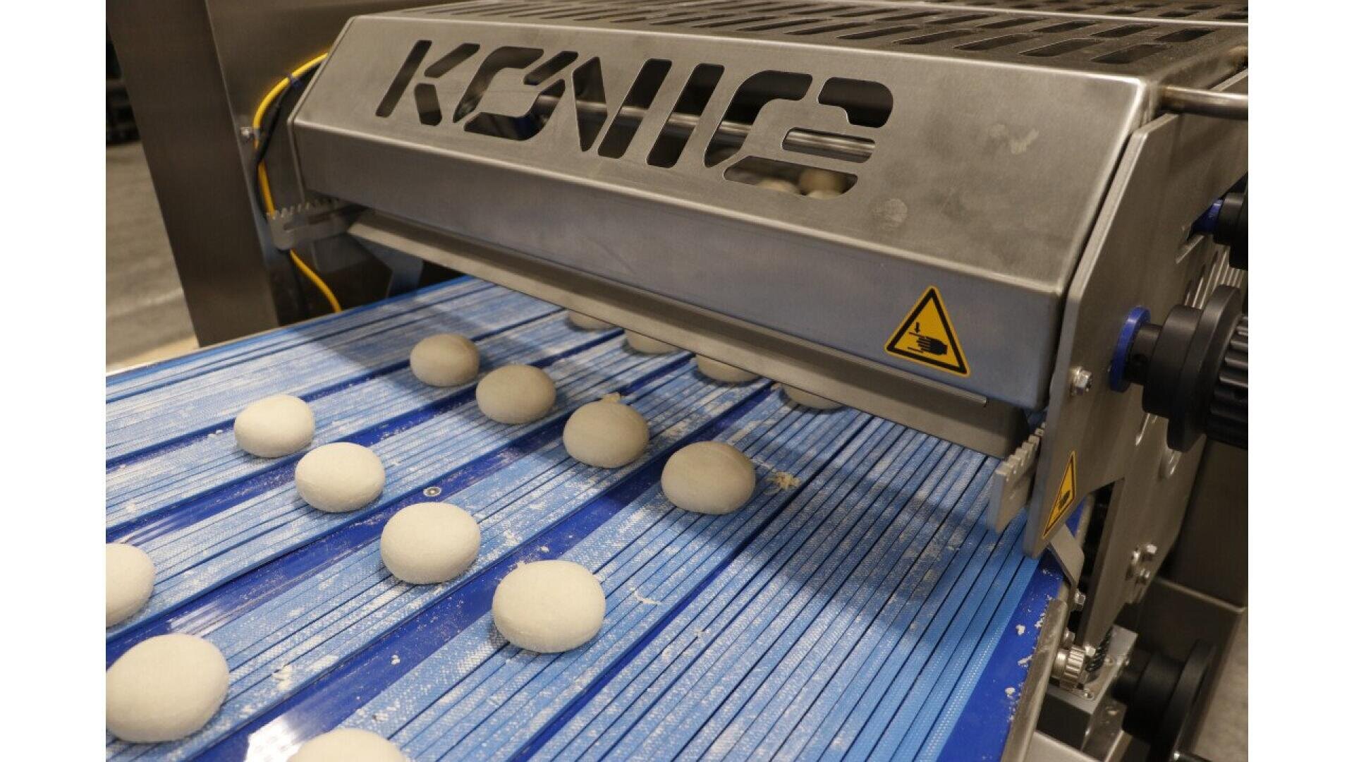 Nahaufnahme einer Teigkugelformmaschine auf einem blauen Förderband mit gleichmäßig verteilten runden Teiglingen, die sich unter einem Metallgerät mit der Aufschrift KONIG bewegen.