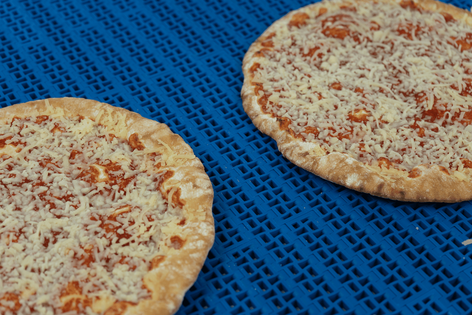 Zwei ungebackene Käsepizzen mit Tomatensoße und geriebenem Käse liegen nebeneinander auf einer blauen, perforierten Oberfläche.
