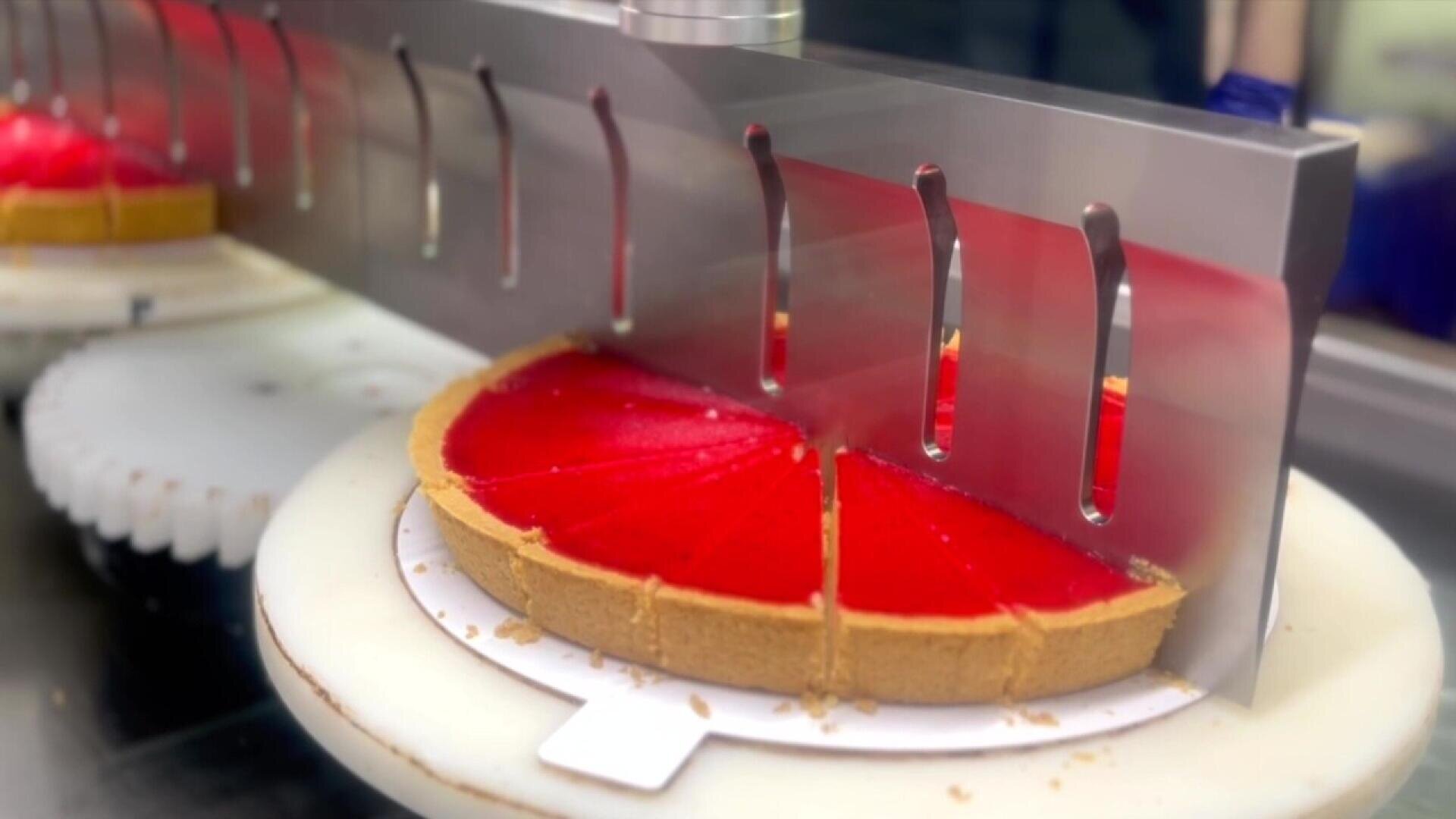 Eine Maschine mit Metallmessern schneidet einen runden roten Kuchen mit Kruste in gleichmäßige Segmente auf einer weißen, rotierenden Plattform.